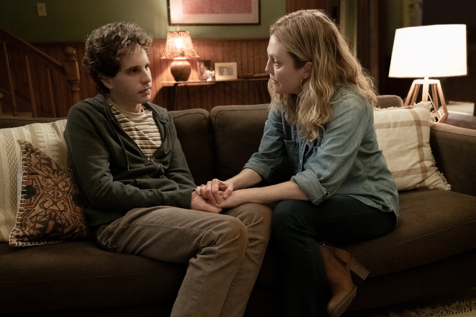 Julianne Moore comforts Ben Platt in the 'Dear Evan Hansen' movie. They sit on a brown couch, Moore's hands clasping Platt's. Moore wears a light blue denim button-up shirt with jeans and heeled booties. Platt wears light brown pants with a striped T-shirt and green zip-up hoodie. 
