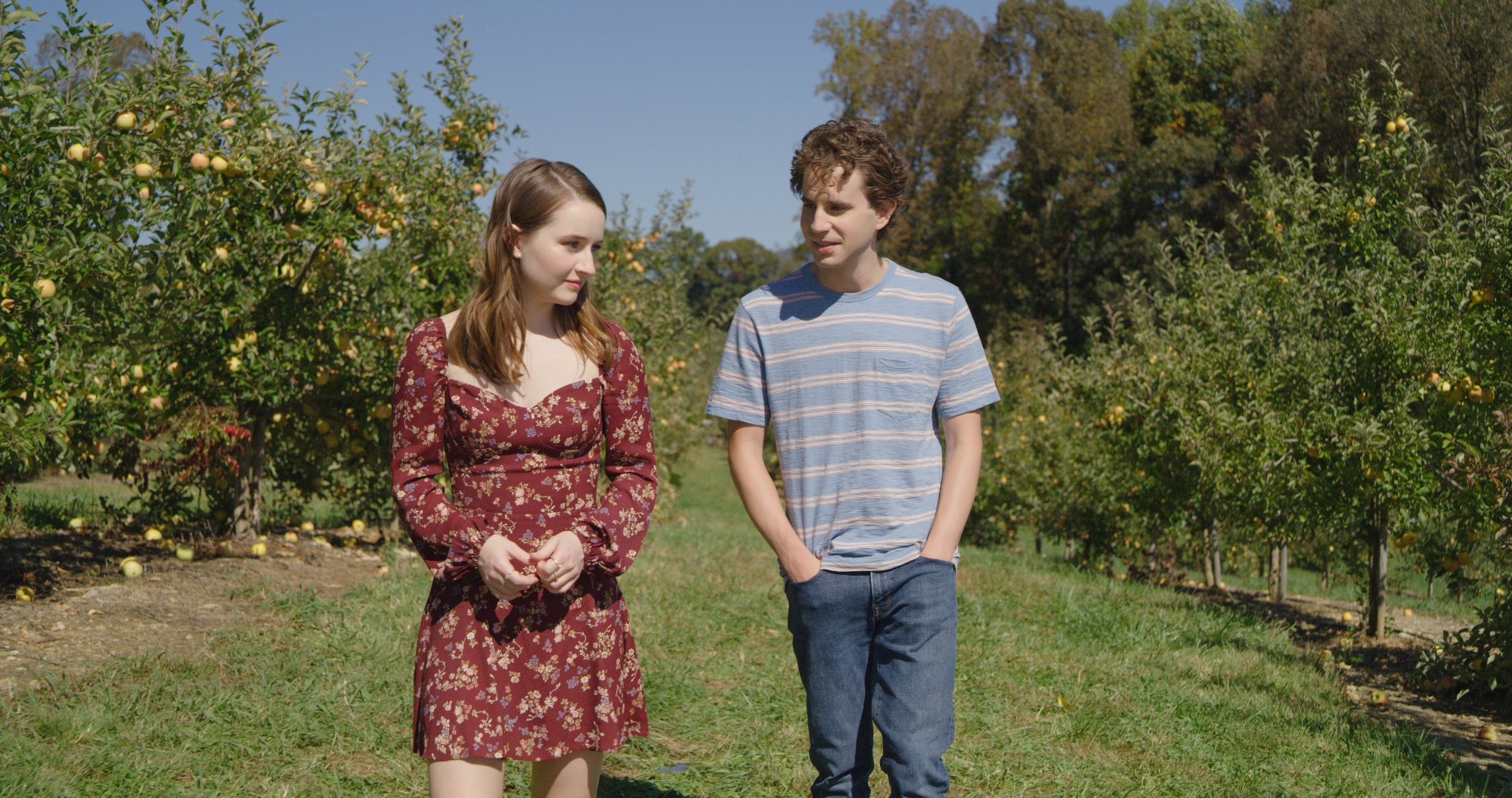 'Dear Evan Hansen' actors Kaitlyn Dever and Ben Platt walking through nature together. Dever wears a burgundy floral dress. Platt wears a blue striped T-shirt and jeans. It's daytime.