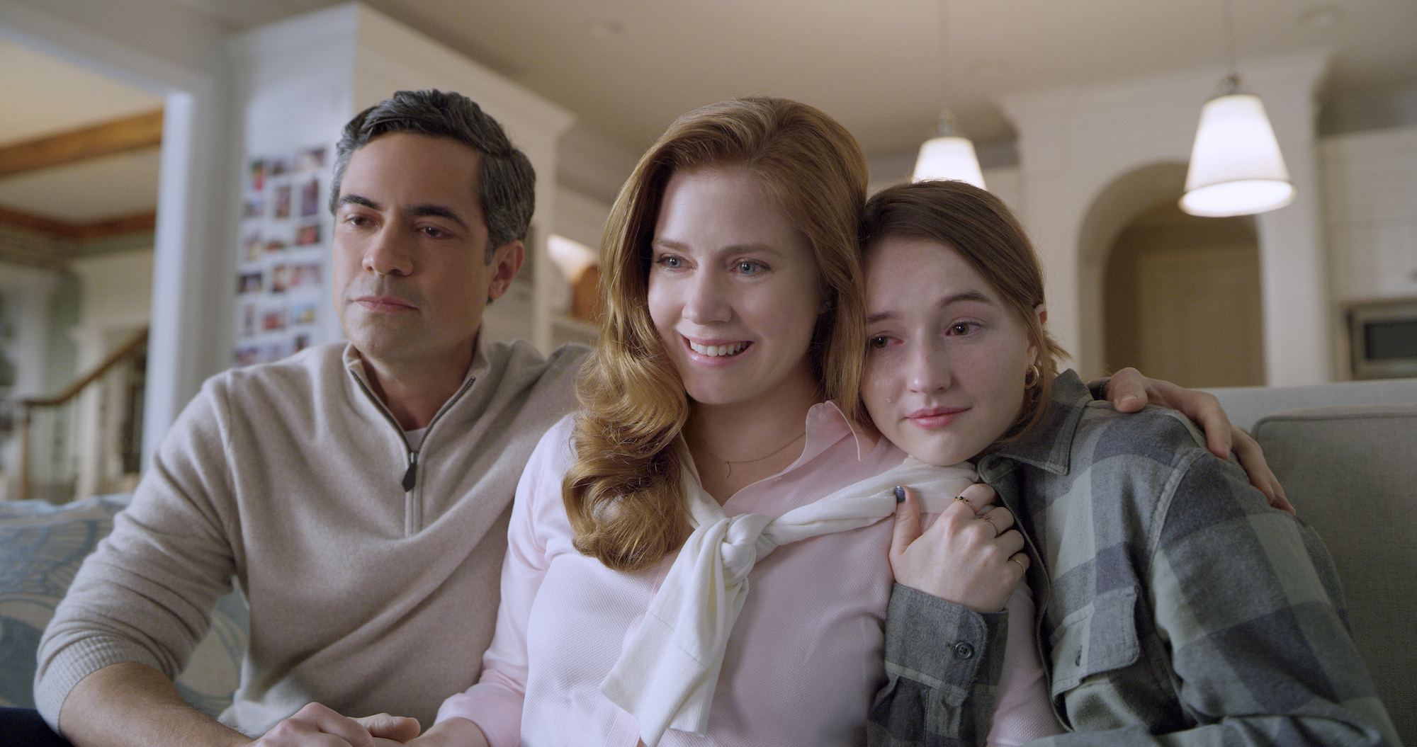 Danny Pino as Larry Mora, Amy Adams as Cynthia Murphy, and Kaitlyn Dever as Zoe Murphy in the 'Dear Evan Hansen' movie. They sit on a couch in a home with their arms around each other. Adams is smiling and all of them have tears in their eyes.