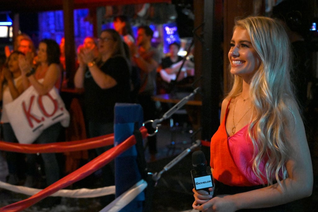 Demi Burnett holds a microphone while participating on 'The Bachelor'. She's wearing a red dress and her long, blonde hair is down.