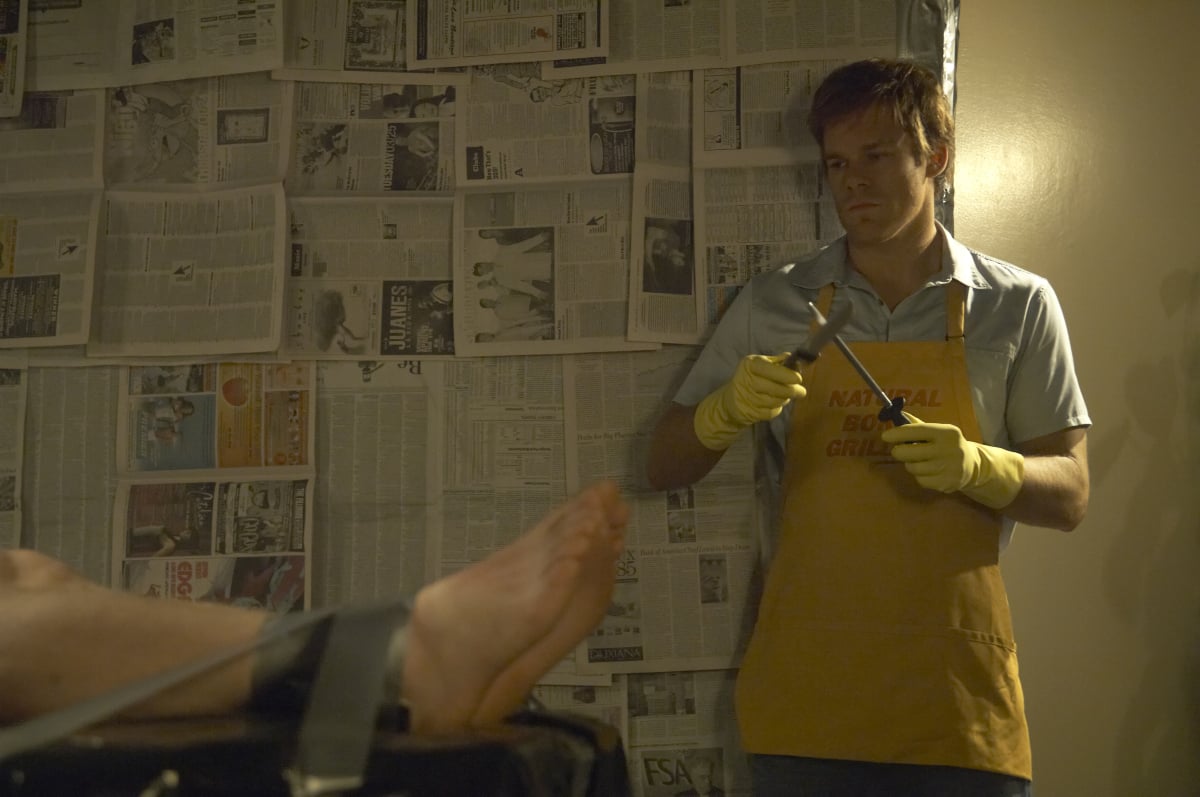 Michael C. Hall as Dexter stands in his kill room sharpening a knife. He is wearing an apron and gloves and looking at the feet of someone wrapped to his kill table.