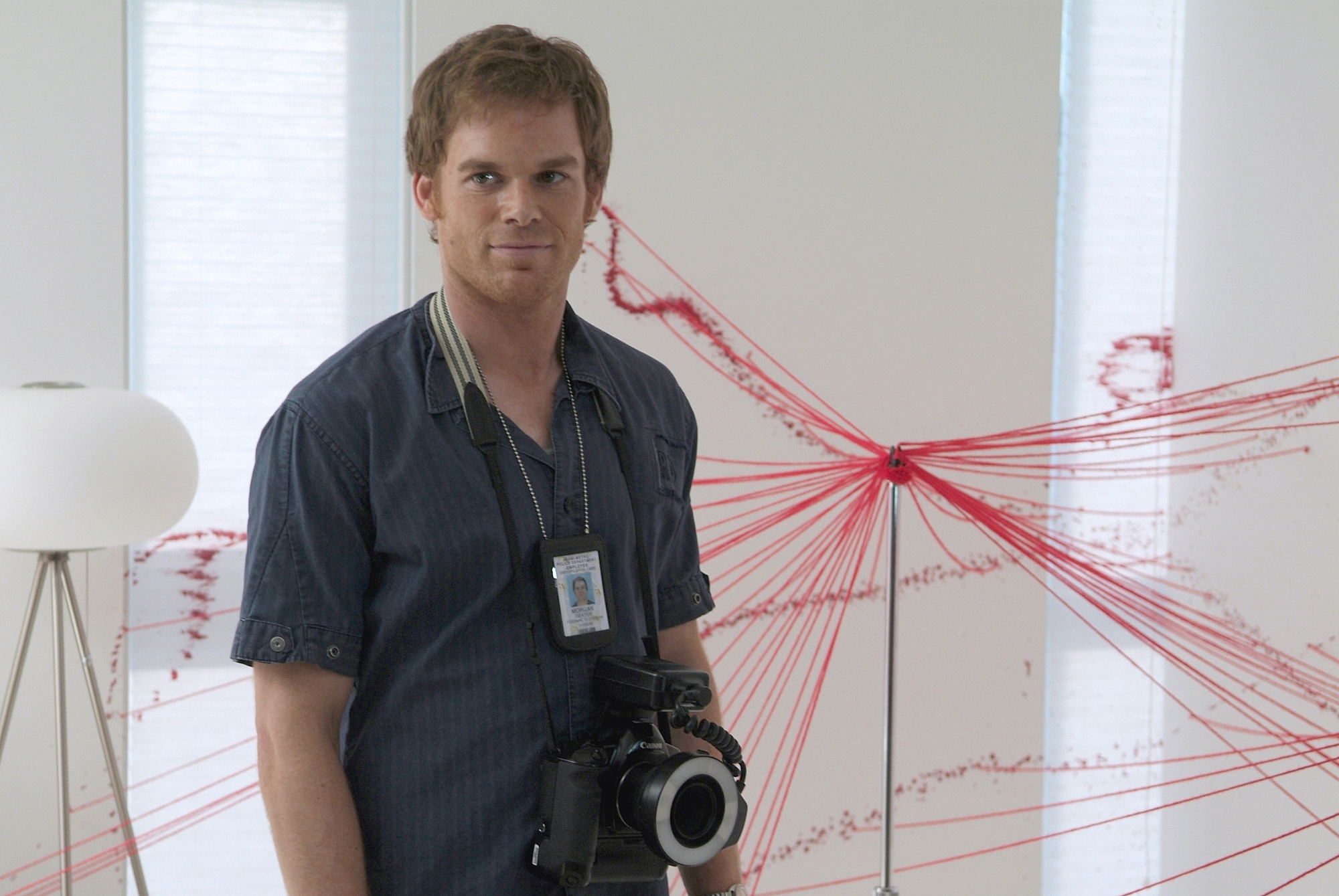 Dexter Morgan stands in front of strings he has attached to various blood splatter patterns. He wears a dark blue collared shirt and a Miami Metro laminate badge. He also has a camera around his neck.