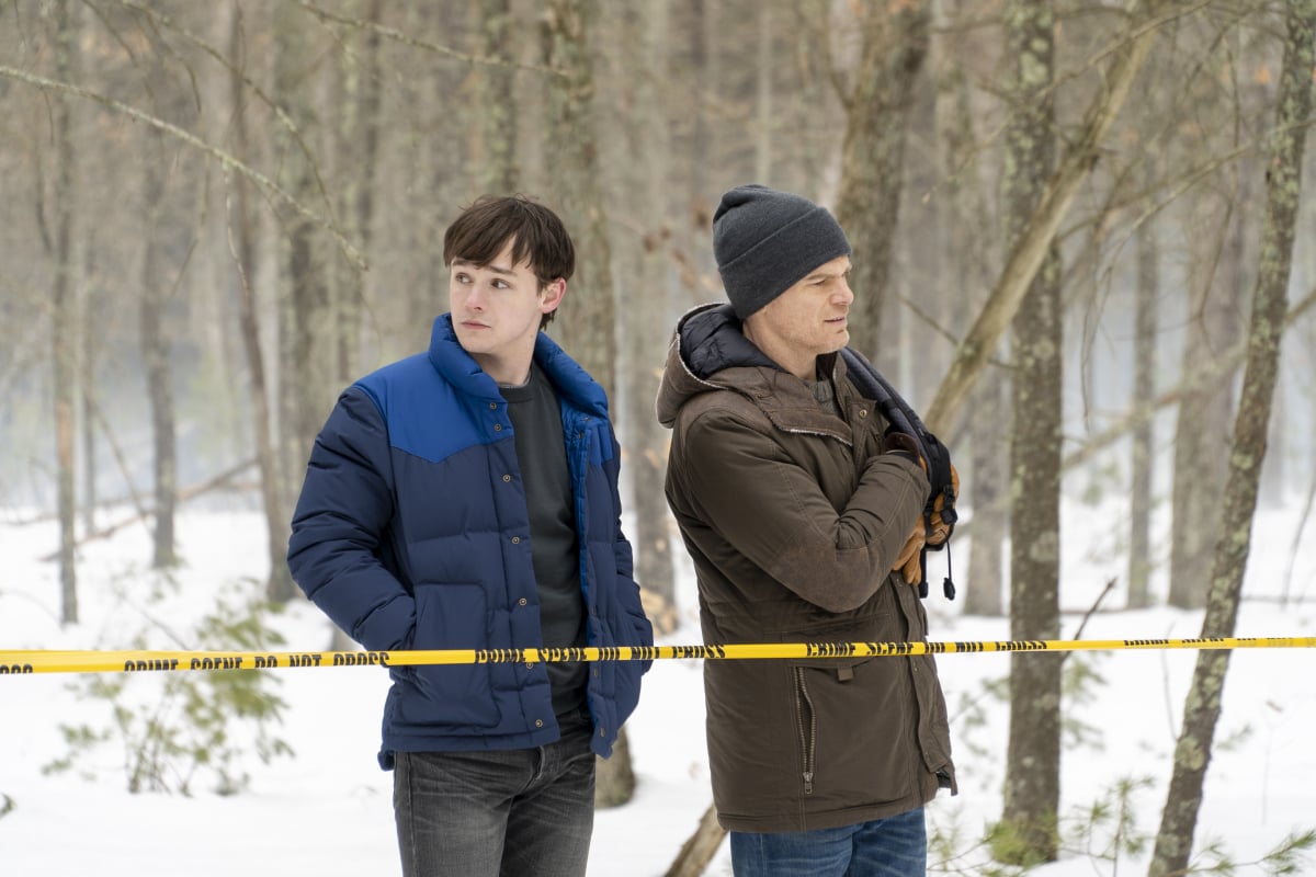 Dexter and Harrison stand behind a taped-off crime scene. It is snowy out, and they are wearing winter coats. Dexter wears a beanie.