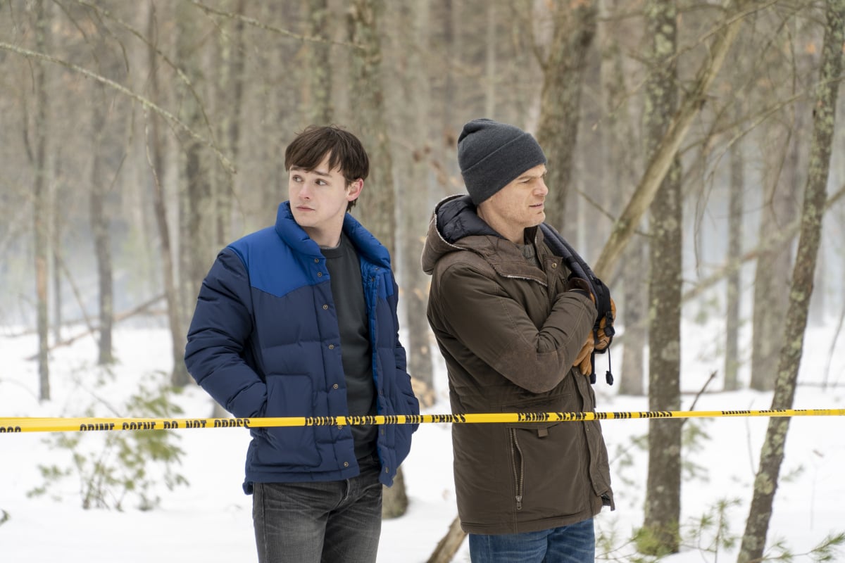 Dexter and Harrison stand behind a taped off crime scene. It is snowy out and they are wearing winter coats. Dexter wars a beanie. 