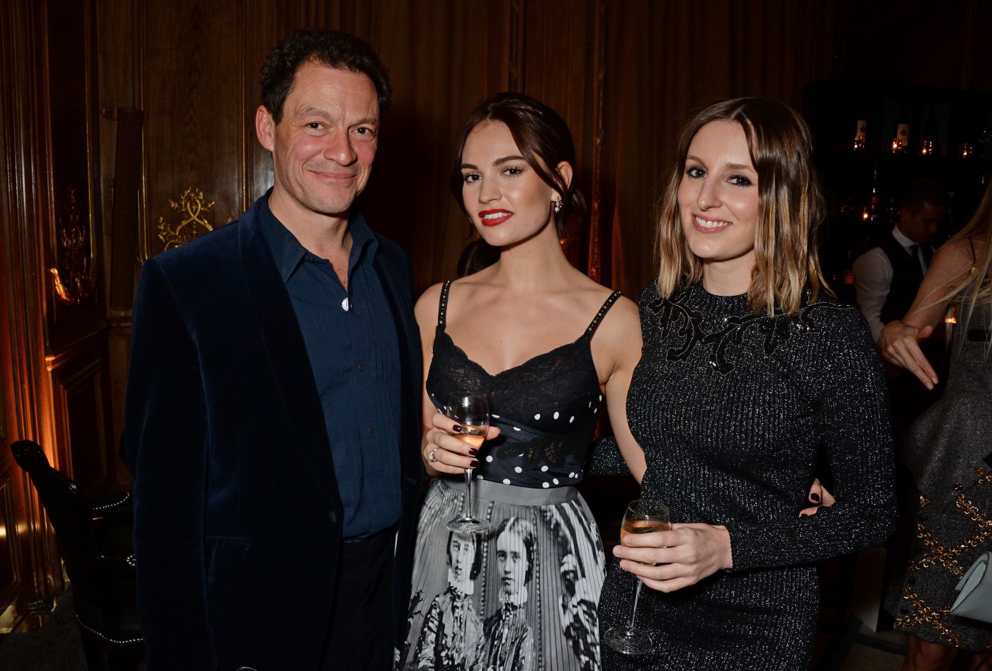 Dominic West, Lily James, Laura Carmichael dressed professionally in front of a dark red curtain.