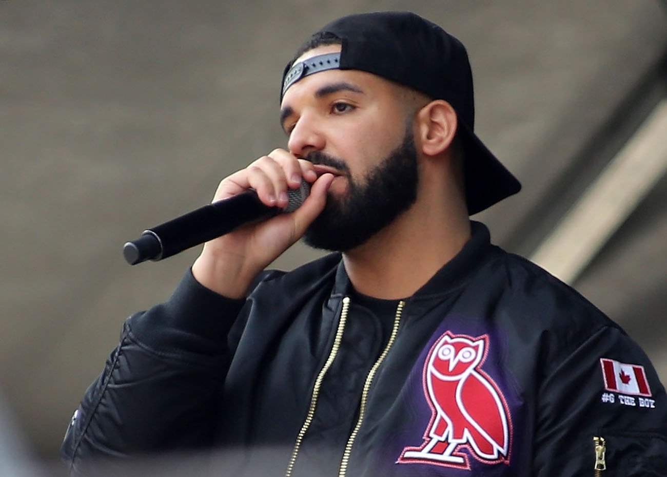 Drake holding a microphone while wearing a black jacket and backward hat.