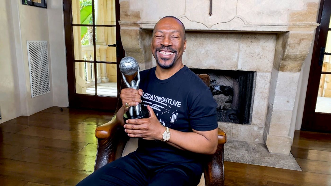 Eddie Murphy dressed in a black t-shirt with white writing in front of a clay colored fireplace.