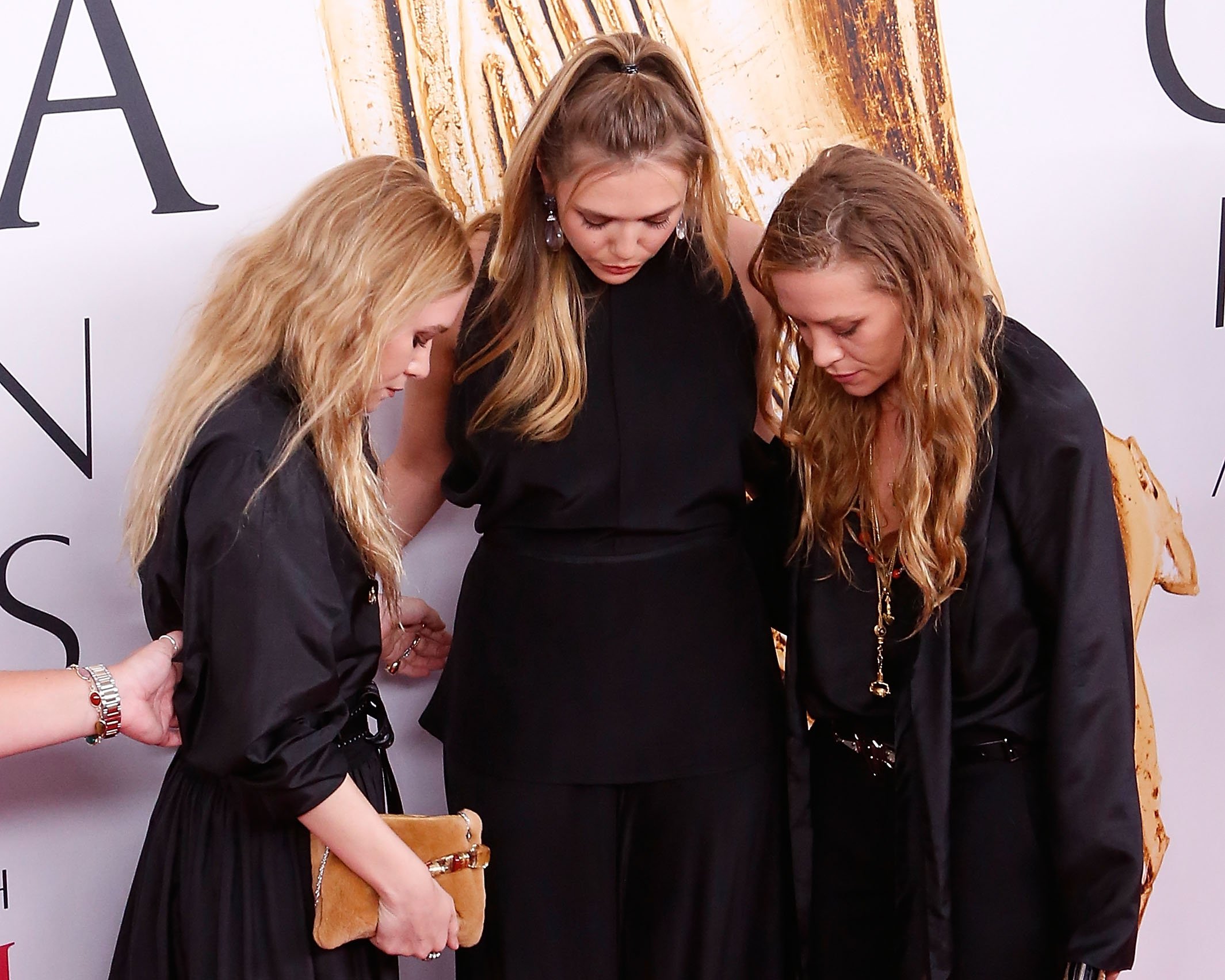 Elizabeth Olsen holding her head down next to Mary-Kate and Ashley Olsen while wearing all black.