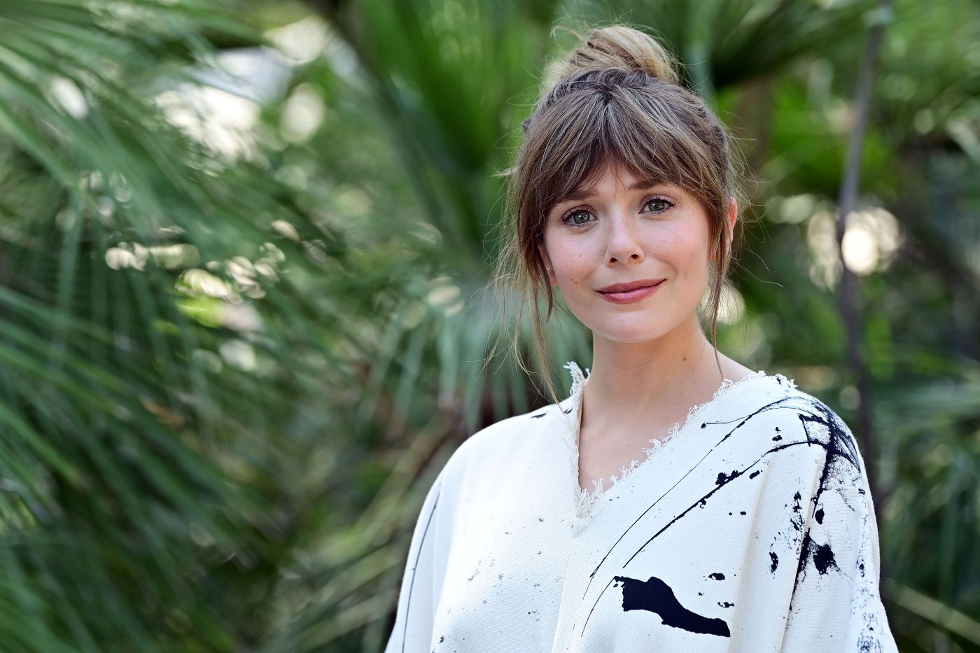 Elizabeth Olsen in a white shirt with black splotches in front of a background with green foliage.