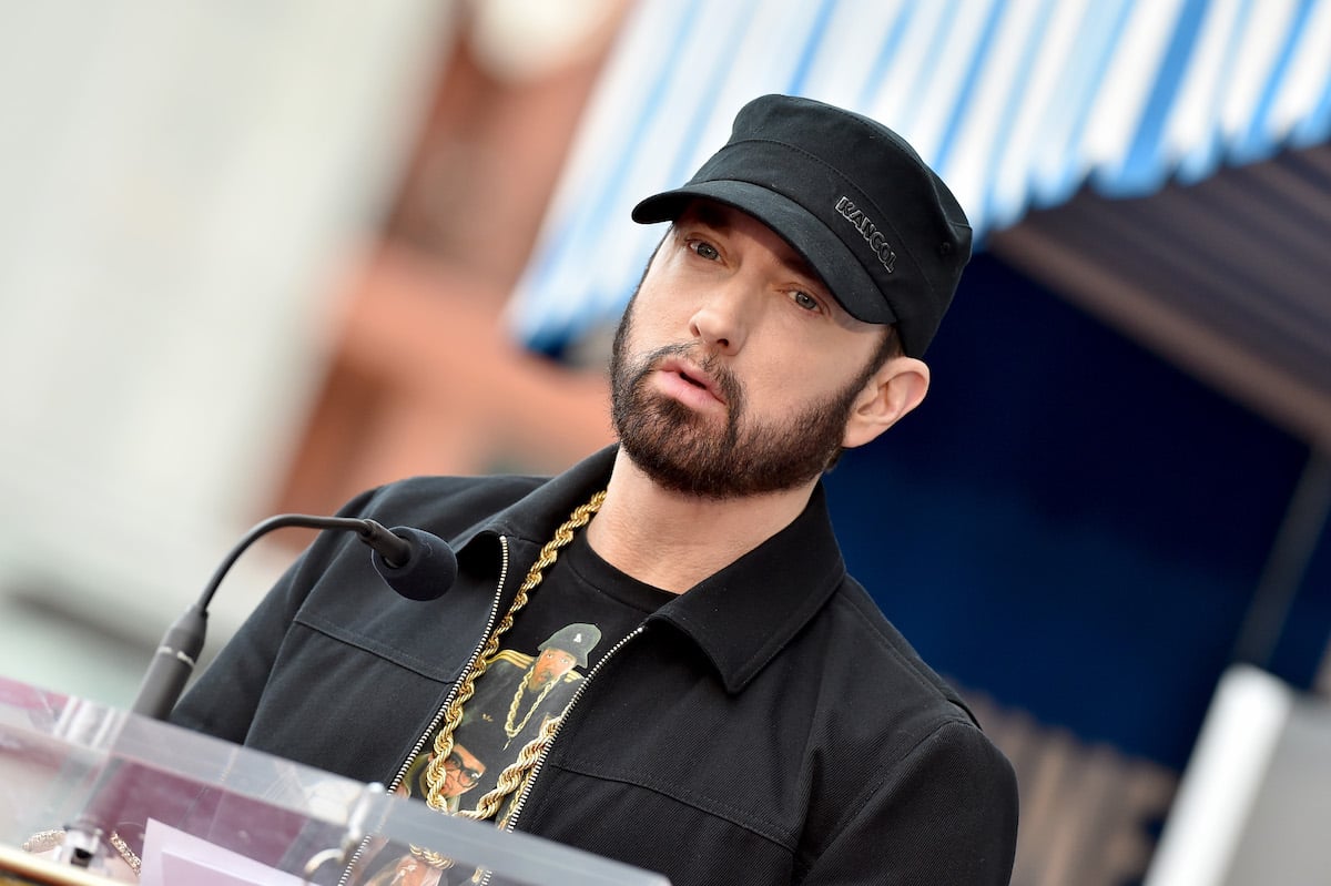 Eminem wears a black outfit and hat on stage speaking into a microphone.