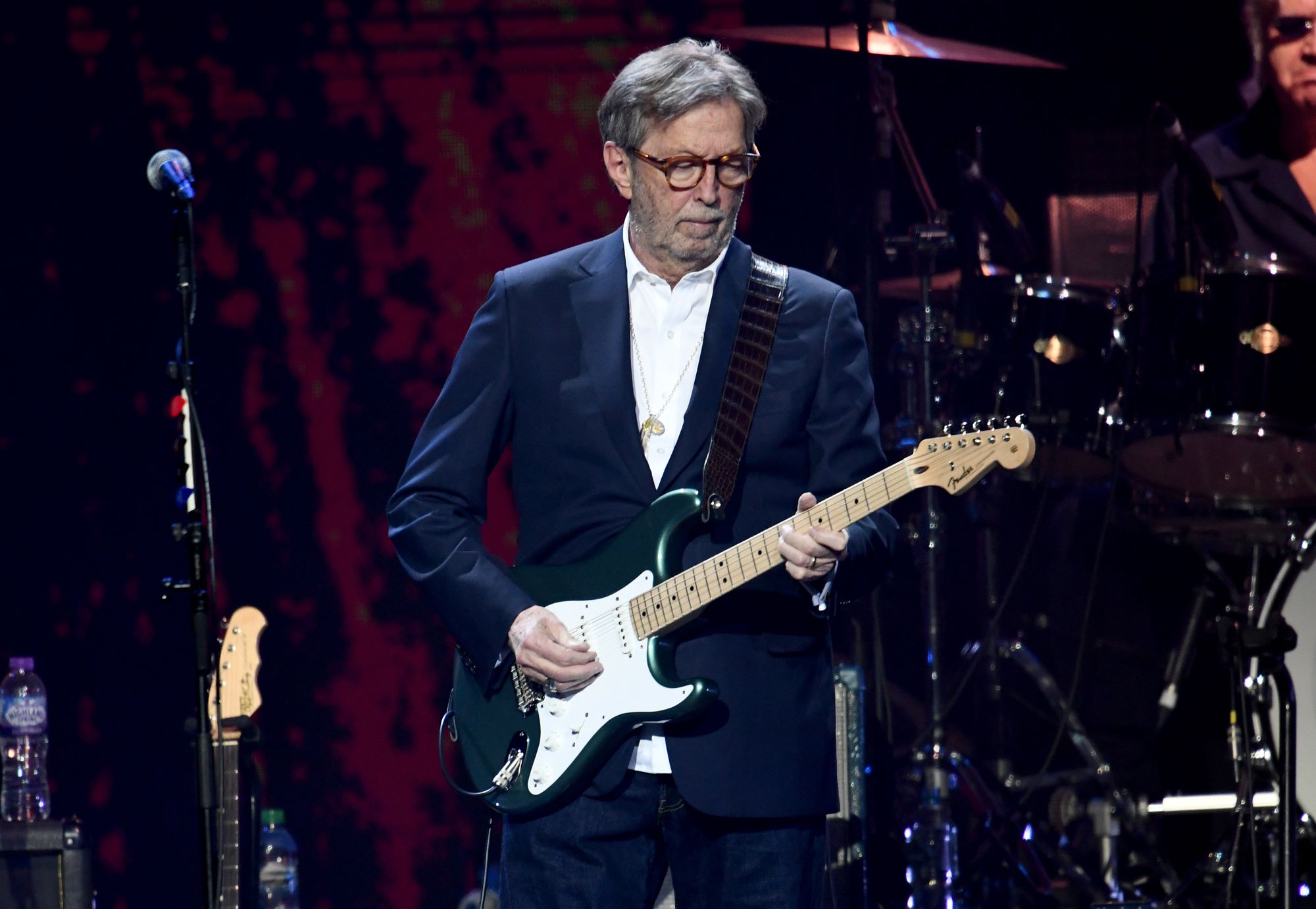 Eric Clapton playing a guitar at one of his concerts wearing a suit in front of a black and red background.