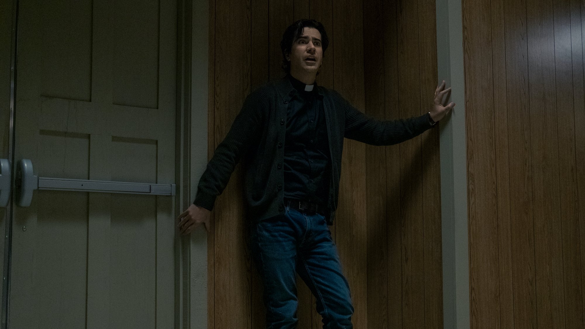 Hamish Linklater as Father Paul crouching in the corner of the Crockett Island rec center in 'Midnight Mass' on Netflix