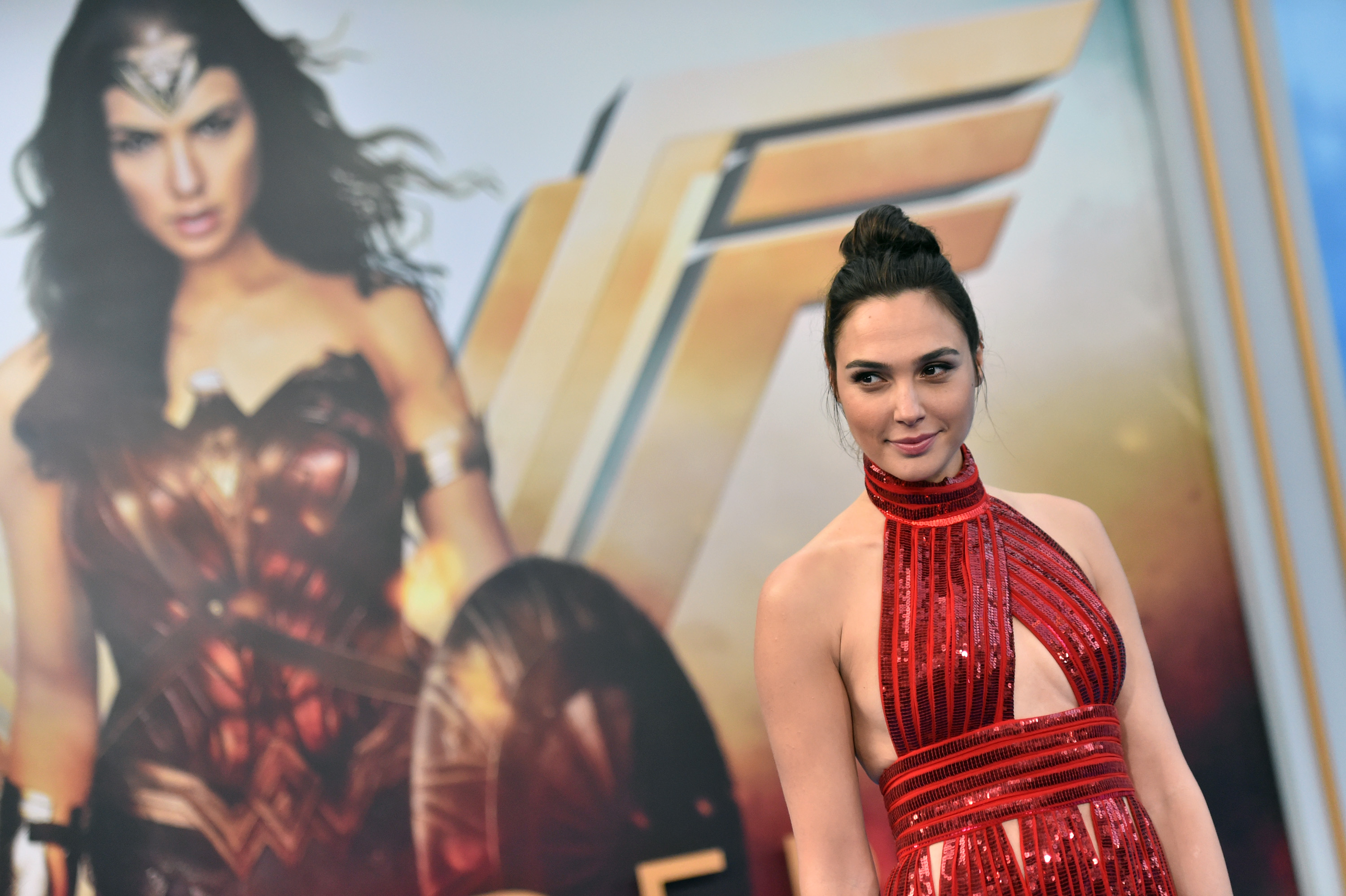 Gal Gadot wears a sparkly red gown and poses in front a picture of her character, Wonder Woman, on the red carpet.