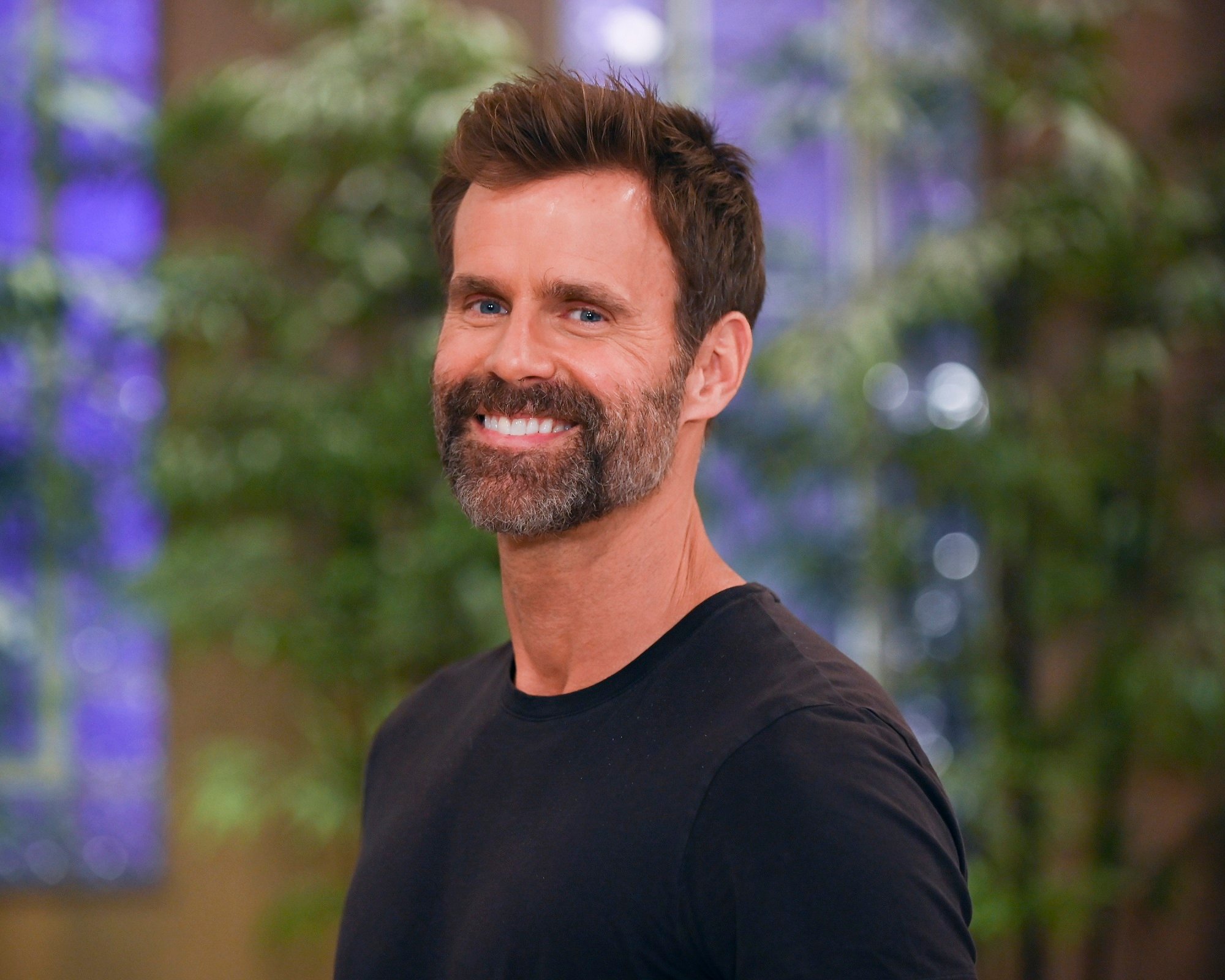 General Hospital star Cameron Mathison, pictured here in a greying beard wearing a black shirt, against a background featuring green plants and blue wallpaper
