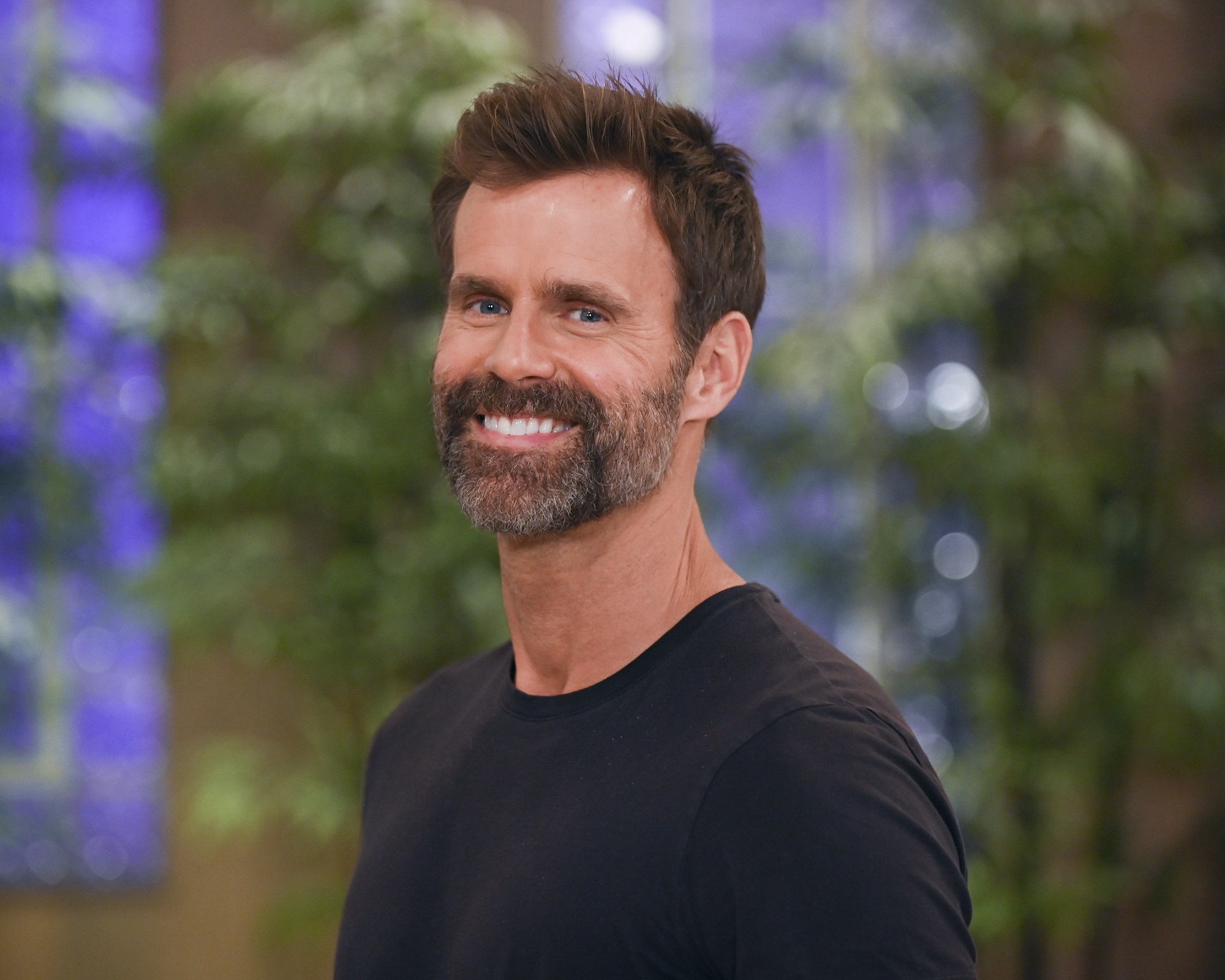 General Hospital star Cameron Mathison, pictured here in a greying beard wearing a black shirt, against a background featuring green plants and blue wallpaper