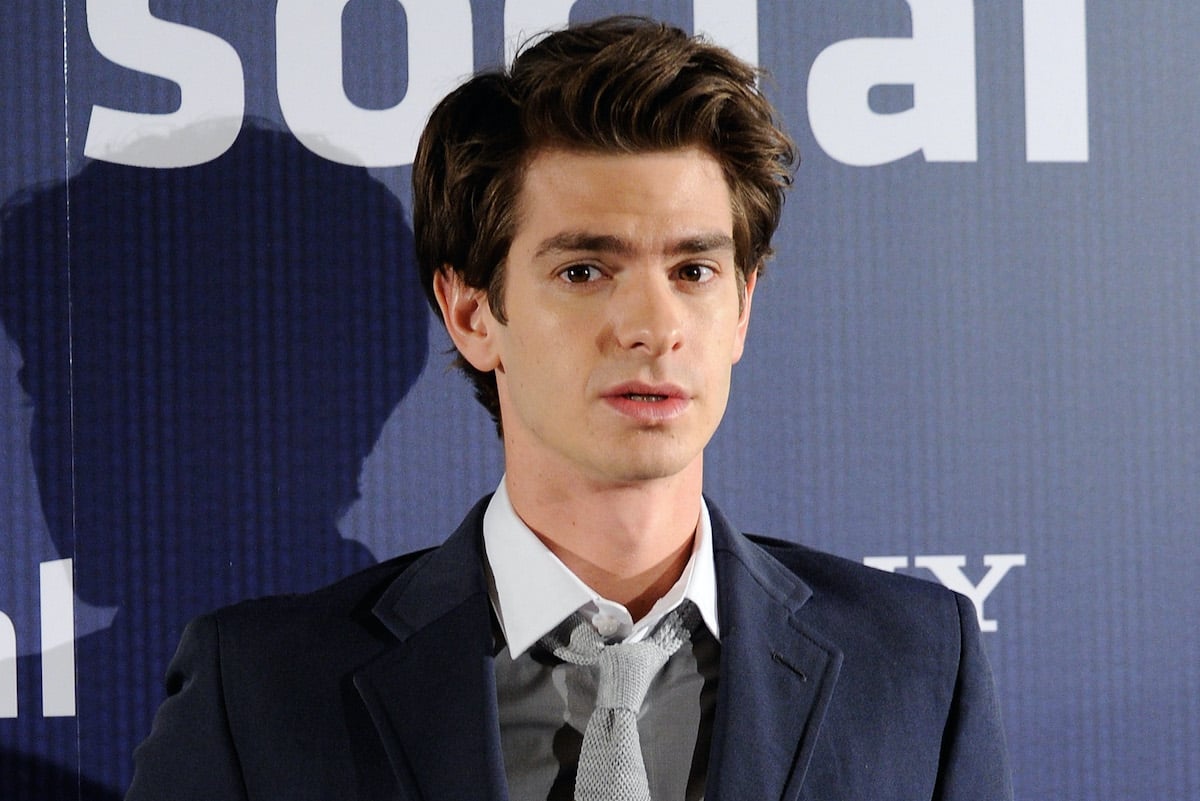 Andrew Garfield at a photocall for 'The Social Network' in 2010. He wears a navy blue suit, grey vest, and light grey tie as he stands in front of a similarly navy blue backdrop that says 'social' and 'Sony' in white lettering. Garfield's 'lawyer up' scene in 'The Social Network' is still one of the most famous from the movie. He revealed what it was like to film it with director David Fincher in a recent interview.