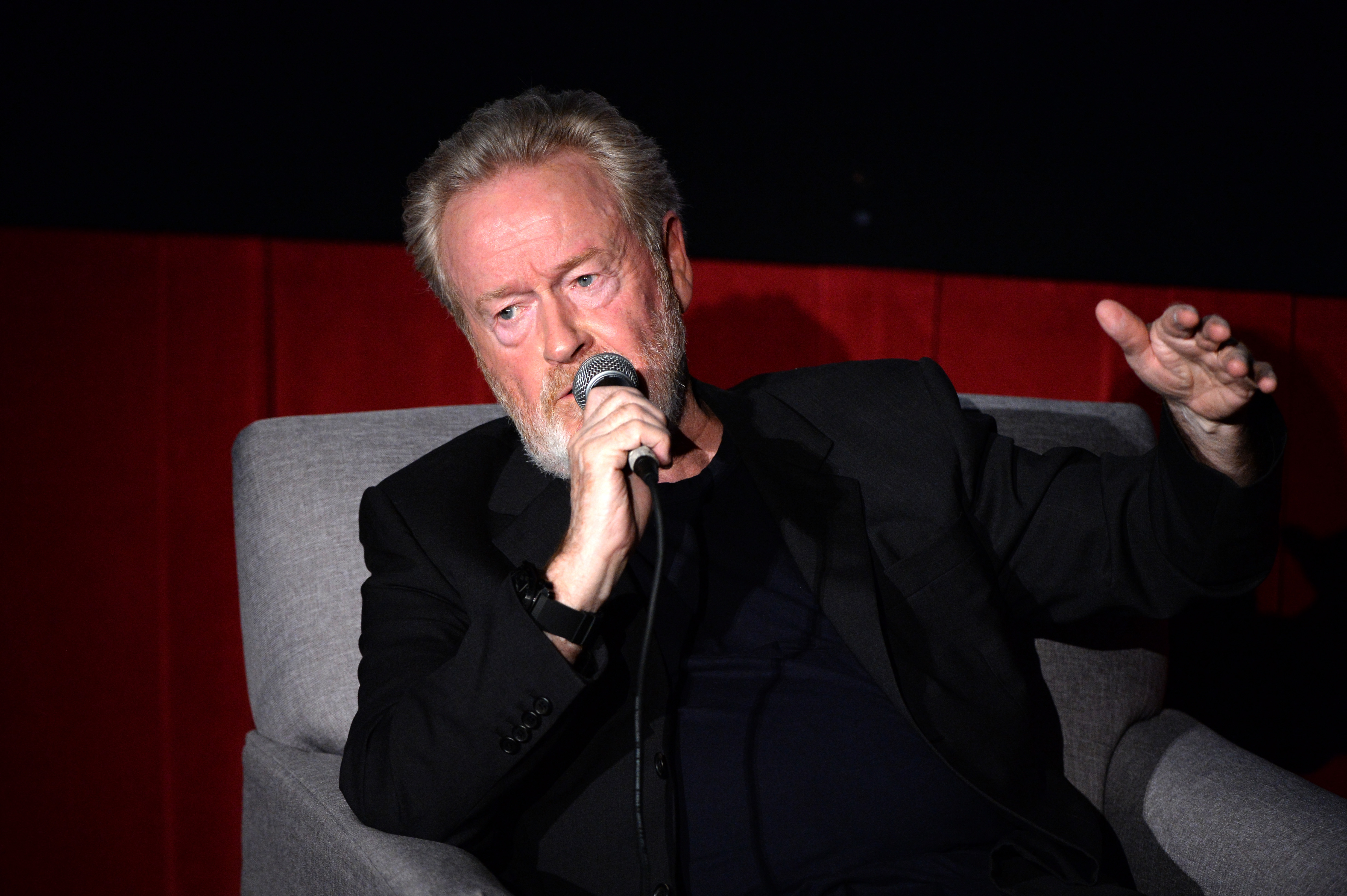 Ridley Scott speaks into a microphone while wearing a black jacket and sitting in a gray chair.