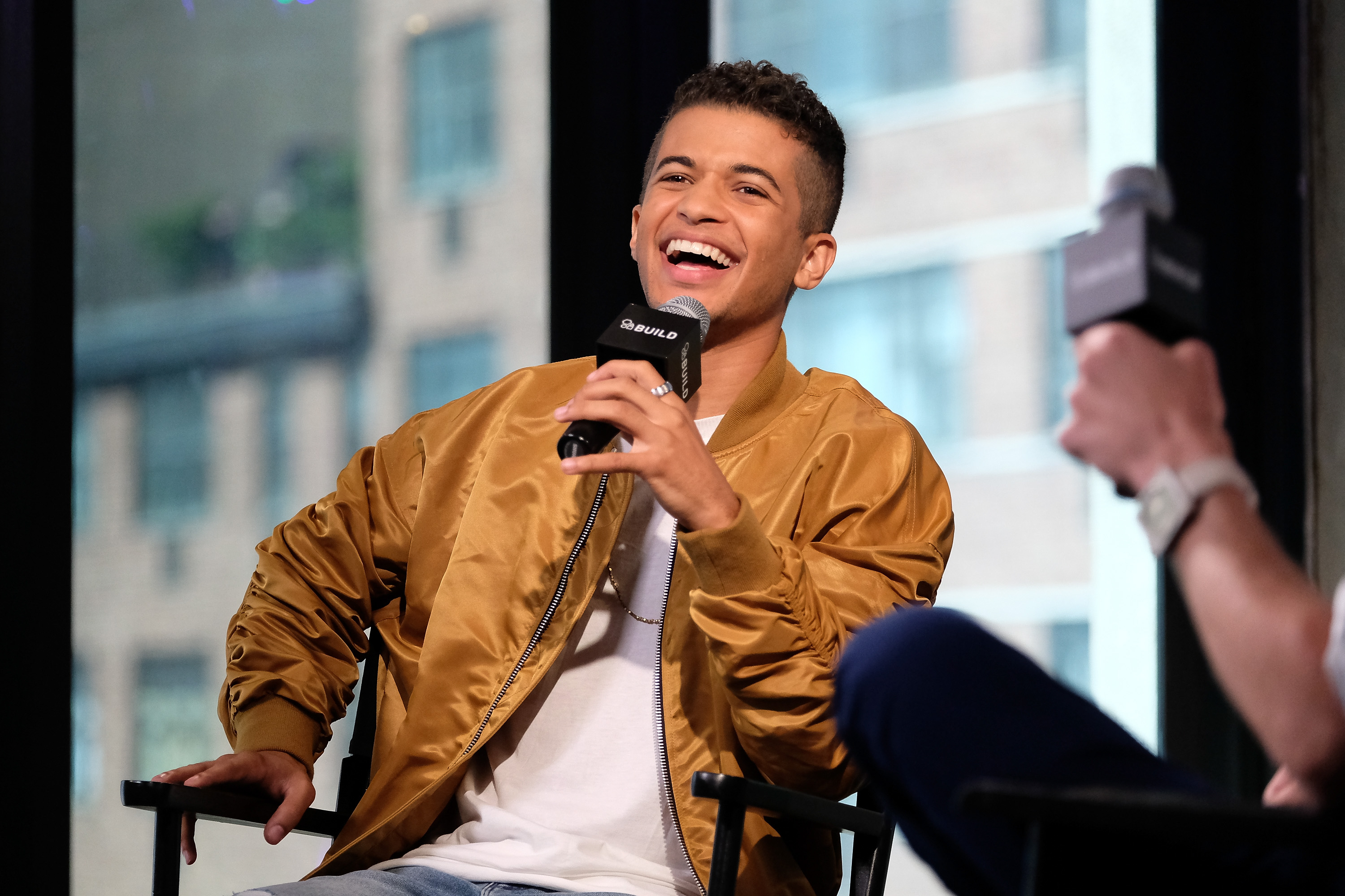 'The Flash' Season 8 star Jordan Fisher wears a brown jacket over a white shirt and laughs into a microphone.