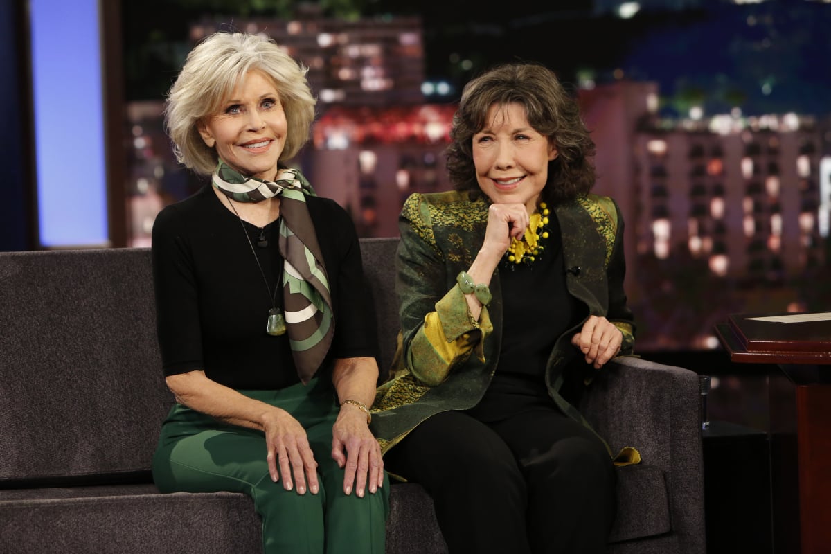 Jane Fonda and Lily Tomlin sit next to each other and smile. Fonda is wearing a black shirt, green pants and a scarf. Tomlin is wearing a green jacket.