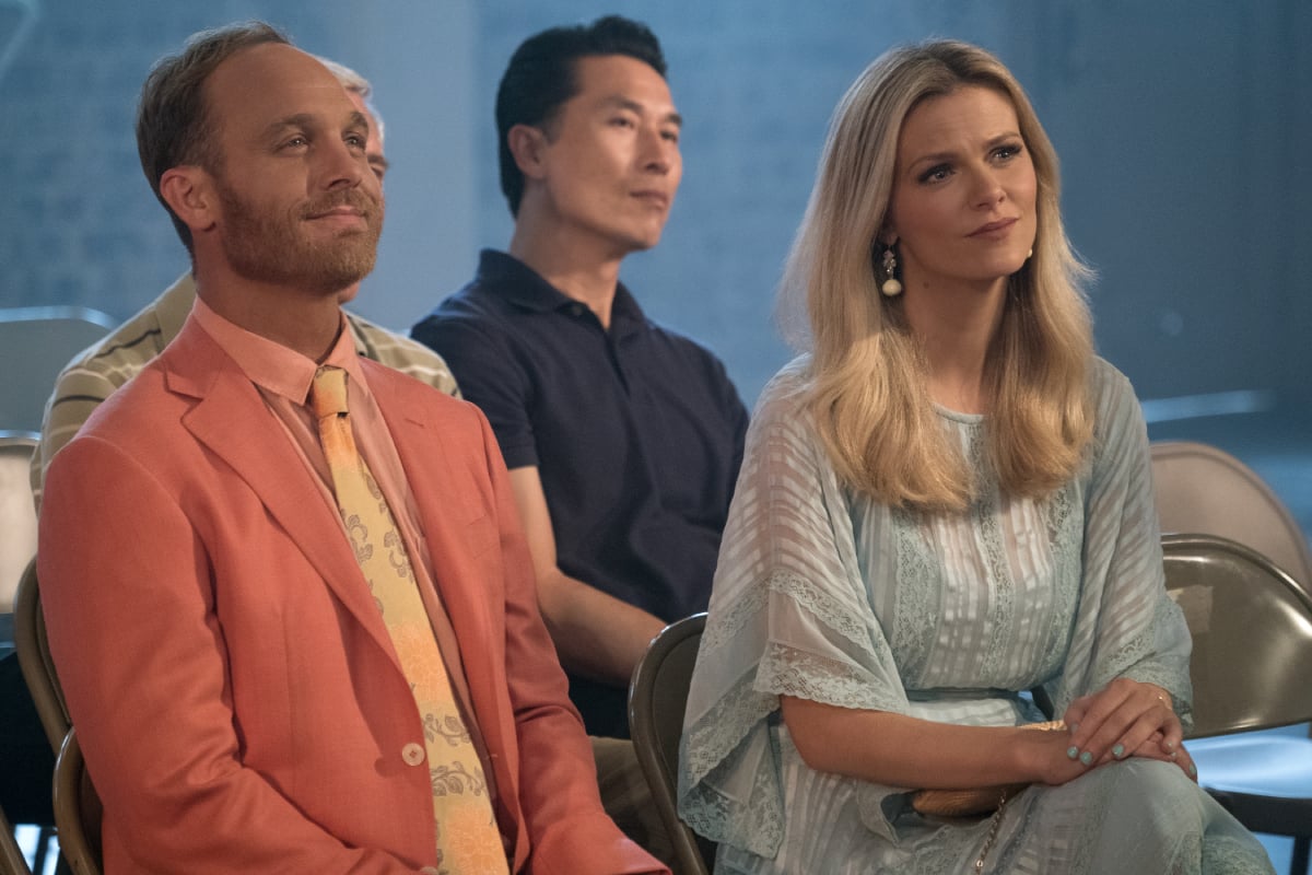 Coyote and Mallory sit beside each other and smile in 'Grace and Frankie.' Mallory is wearing a flowy blue dress and Coyote is wearing a salmon-colored suit and yellow, patterned tie.