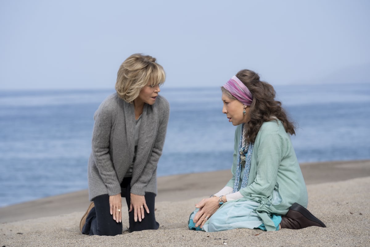 Grace and Frankie kneel sit in the sand on the beach.