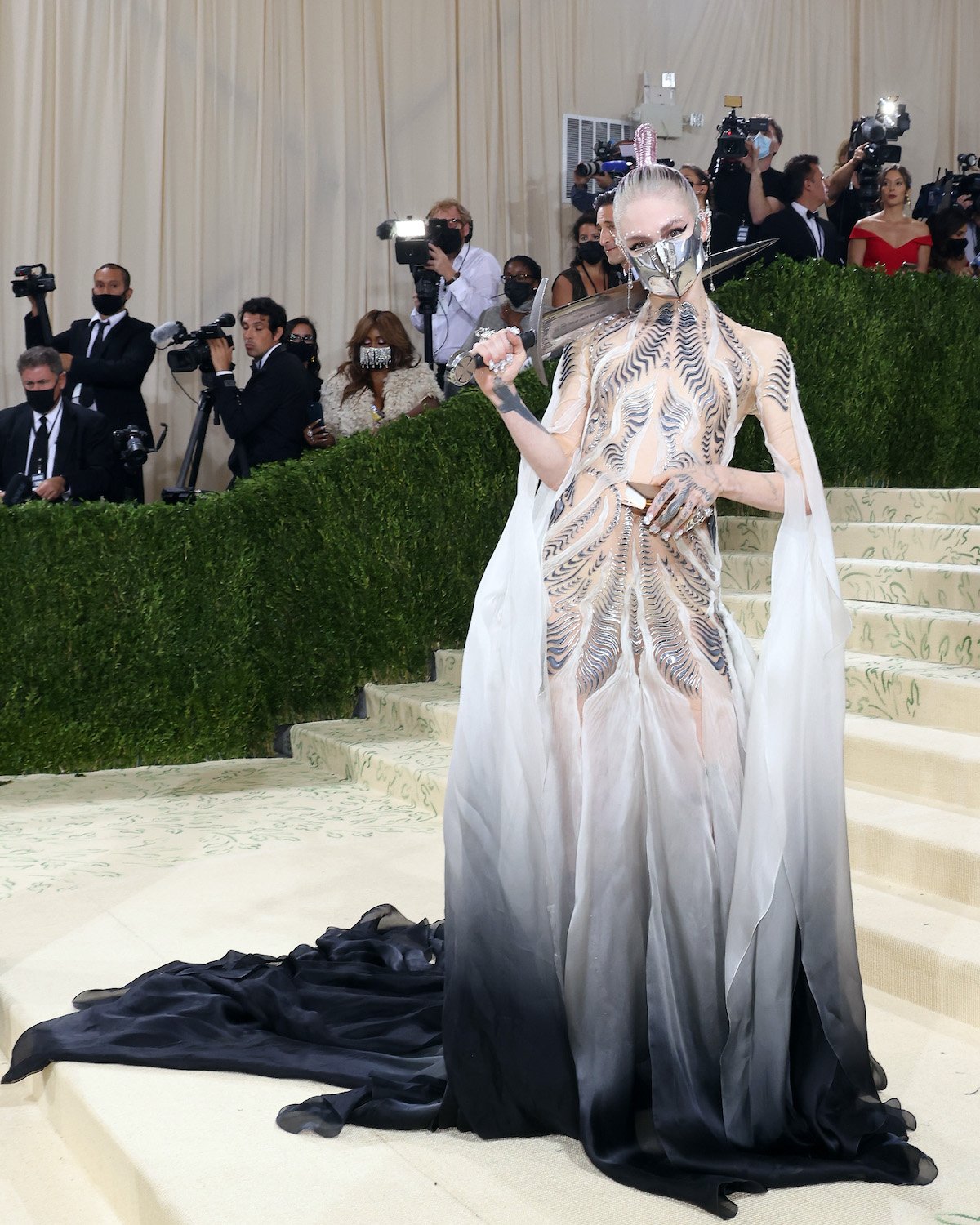 Grimes wears a white to black ombre gown with metallic accents at the 2021 Met Gala. She is holding a sword and a book as part of her look.