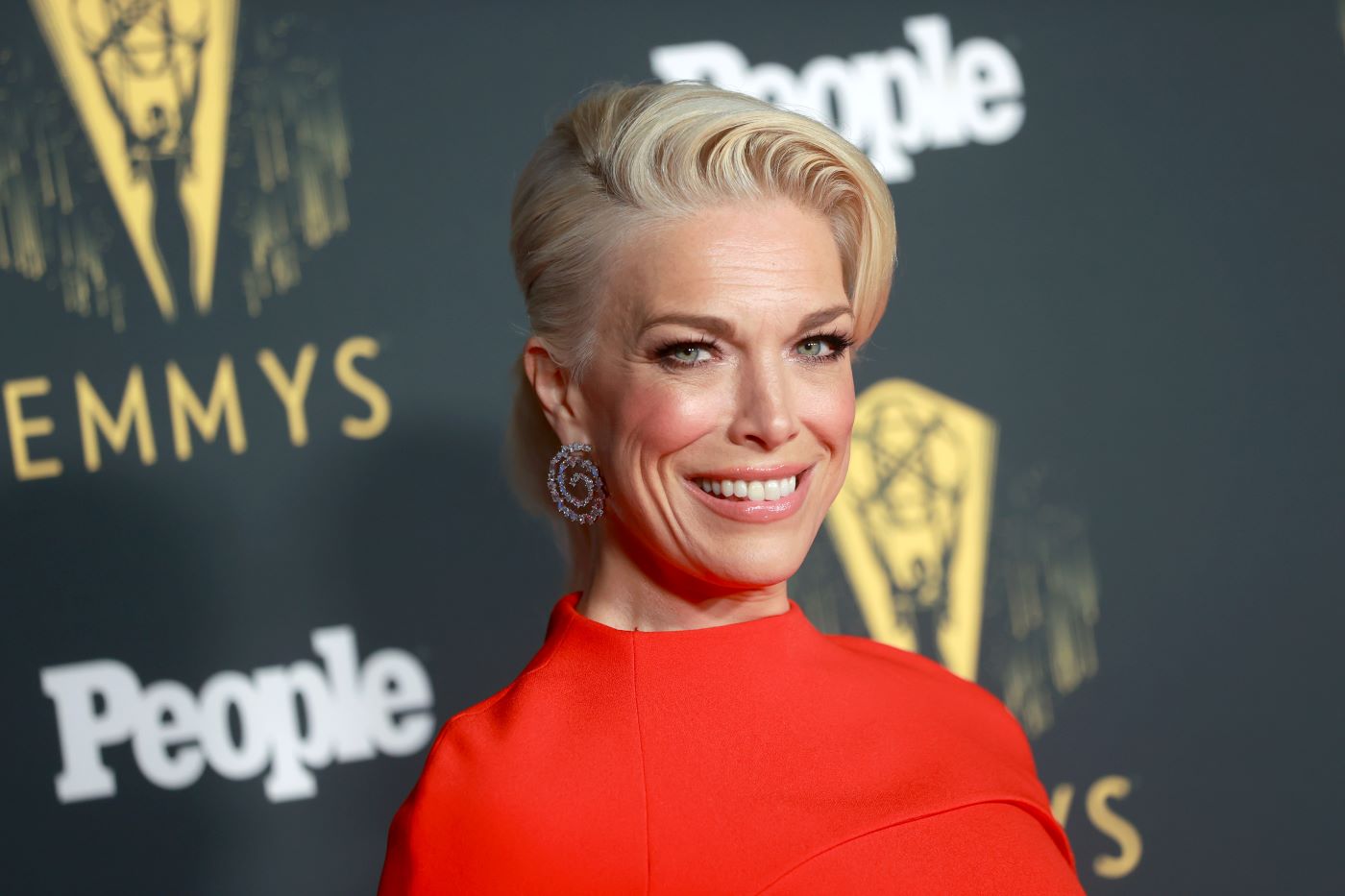 Hannah Waddingham from 'Ted Lasso' wearing a red top in front of a black gold and white background.