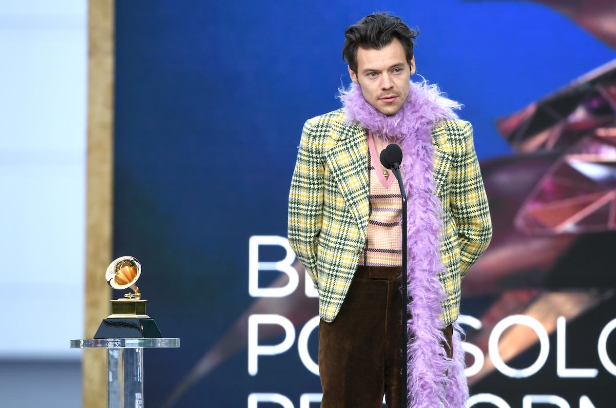 Harry Styles wears a feather boa while speaking into a microphone on stage at an event.