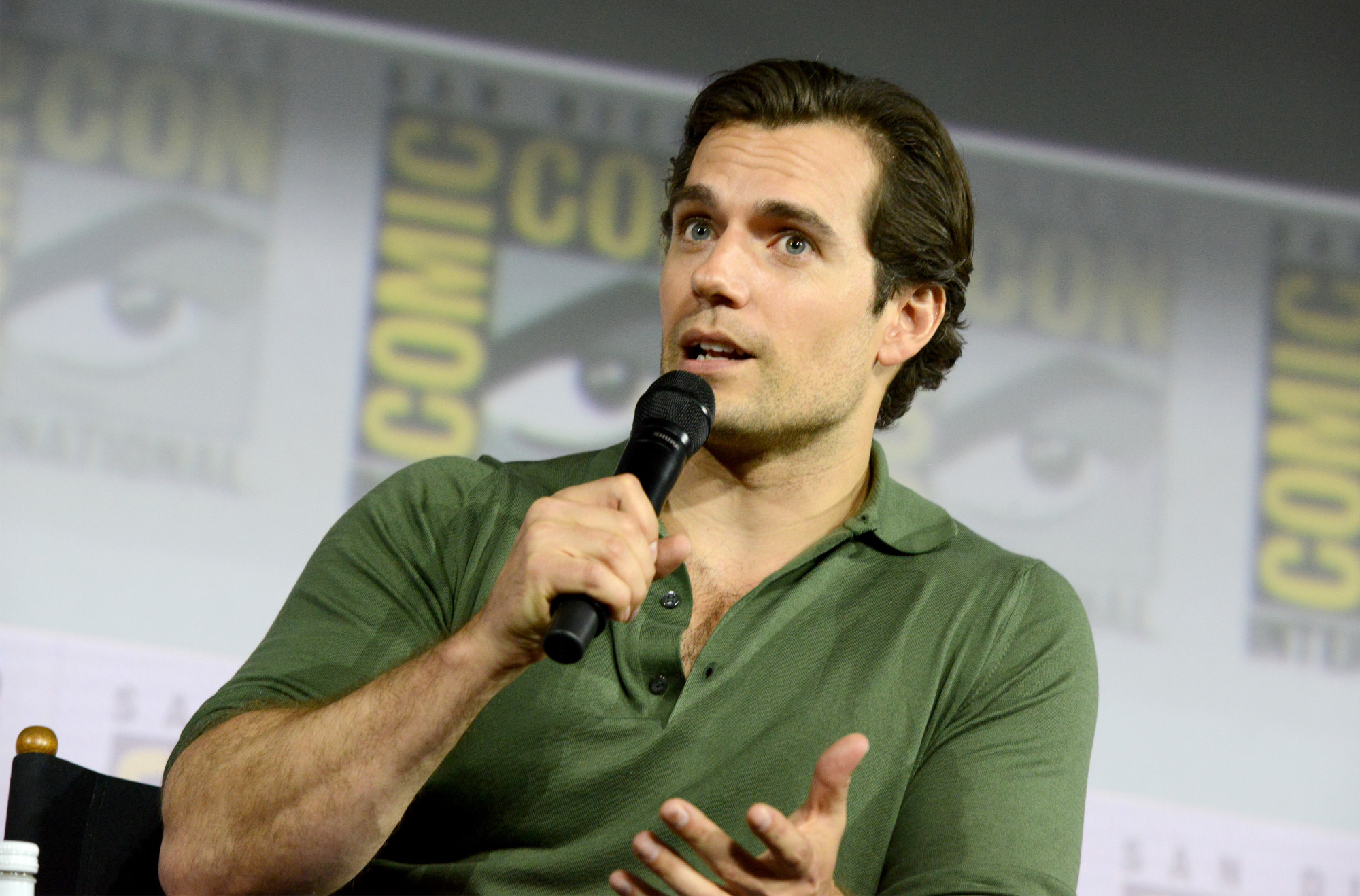 Superman star Henry Cavill during a Comic-Con panel for 'The Witcher.' The actor is wearing a green polo shirt and holding a microphone. He's speaking into it, and a Comic-Con wall is behind him.
