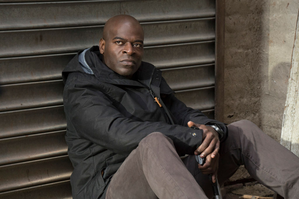 Hisham Tawfiq as Dembe Zuma sits with his back against a wall with an angry look on his face. He's dressed in a puffy black coat and grey pants.