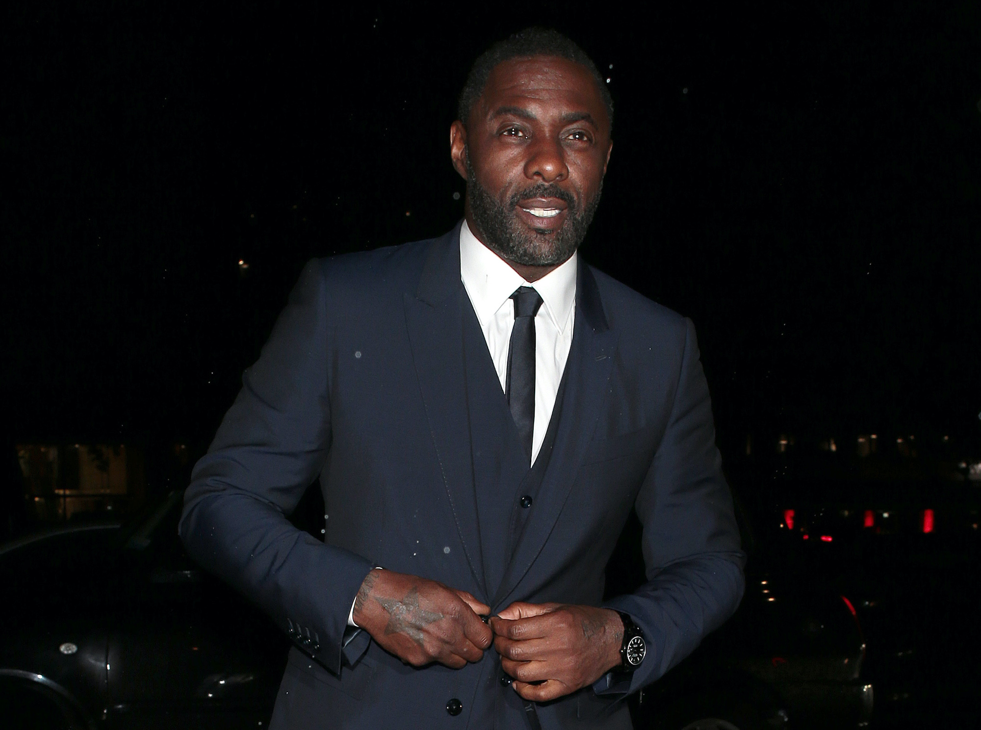 Idris Elba attending Bond: No Time To Die world premiere after parties on Sept. 28, 2021 in London. He walks through a parking lot at night wearing a navy blue suit. He buttons the suit jacket as he walks through the lot. It's raining slightly and there are red car lights behind him.