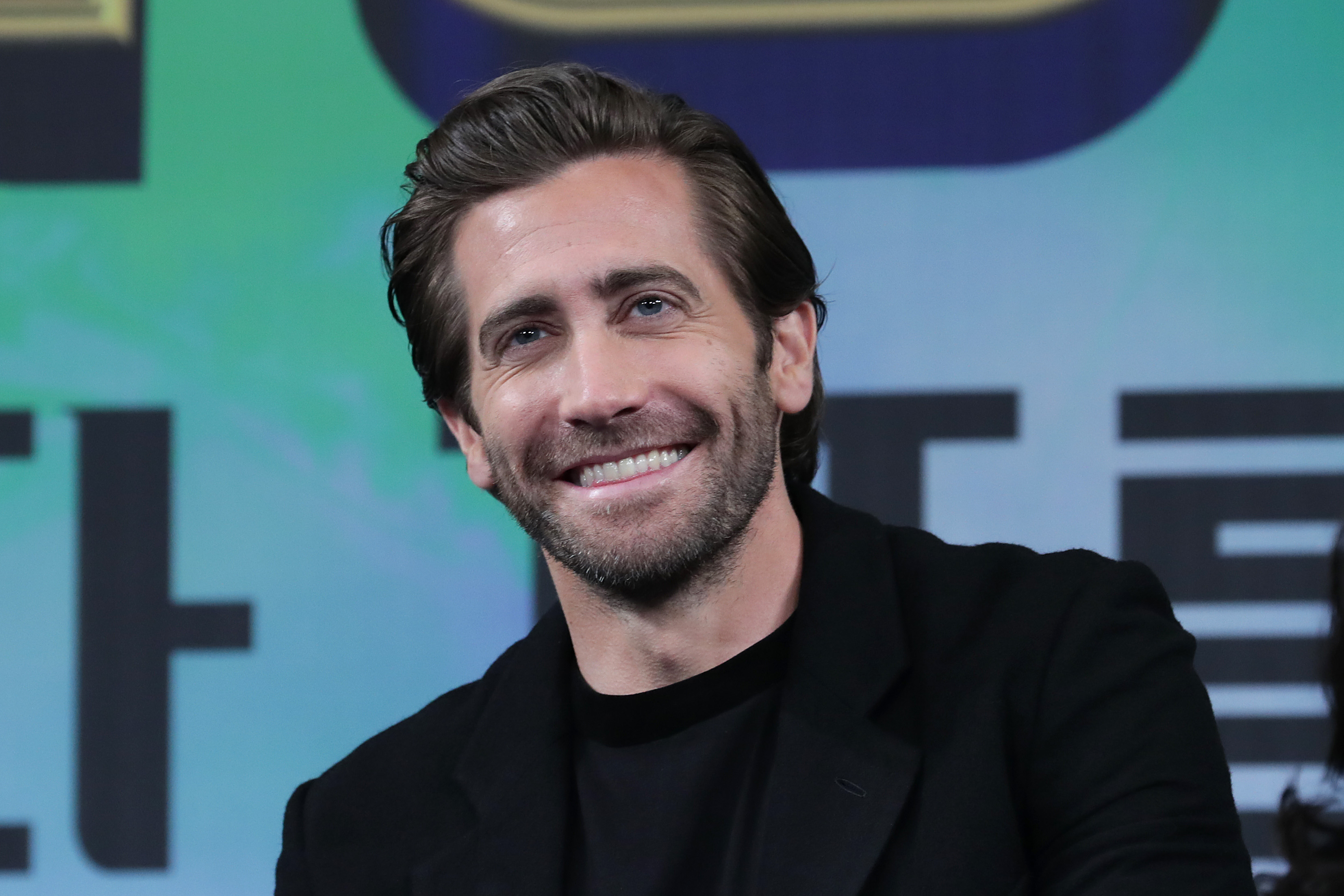 Actor Jake Gyllenhaal, in a black t-shirt and sweater, smiling while at a press conference promoting 'Spier-Man: Far from Home' in Seoul, South Korea in 2019.