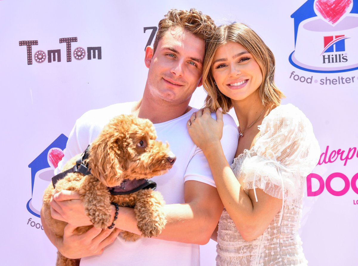 James Kennedy and Raquel Leviss pose together smiling and holding their dog Graham at an event.