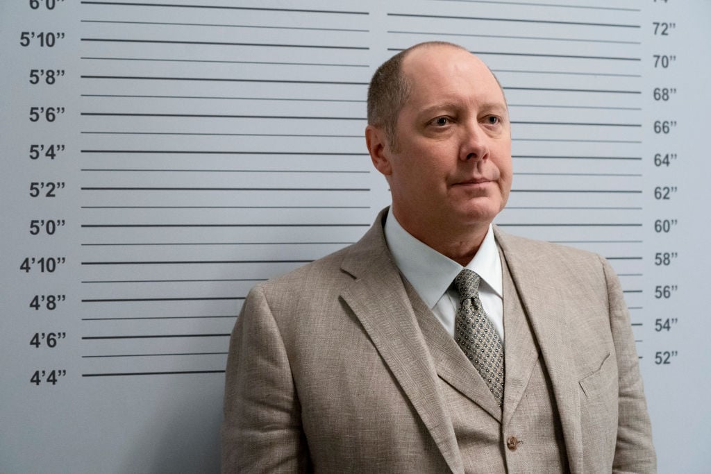 James Spader as Raymond 'Red' Reddington stands in a police lineup for a photo. He's wearing a tan suit.