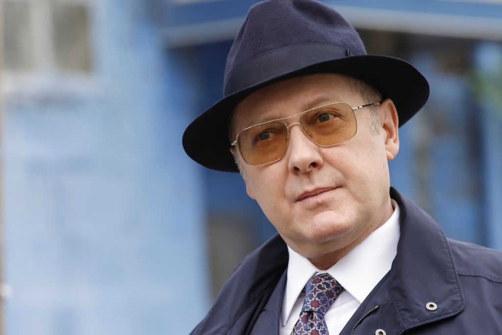 James Spader as Raymond 'Red' Reddington is wearing a navy fedora and matching coat with sunglasses.
