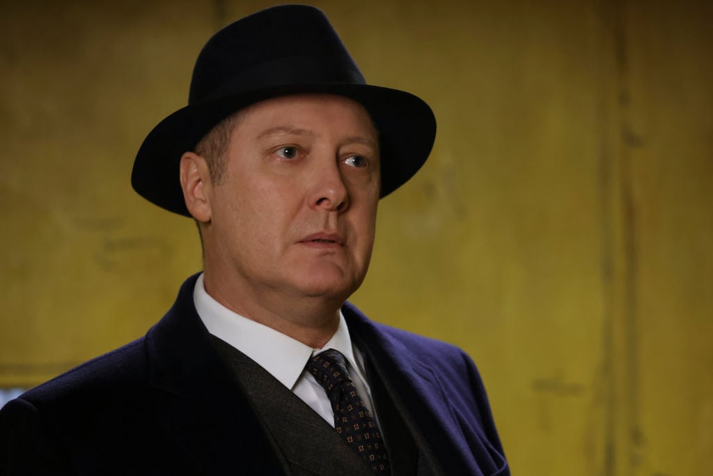 James Spader as Raymond 'Red' Reddington stands in a navy suit with matching fedora and a confused look on his face.