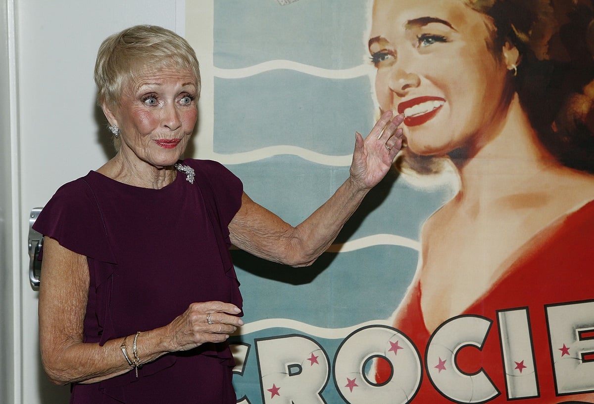Jane Powell amiles and looks at the camera while points to a poster to one of her movies.