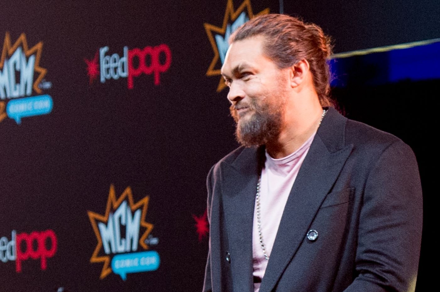Jason Momoa dressed in a black suit with a lavender shirt with a black background with red, blue, orange, and white writing.