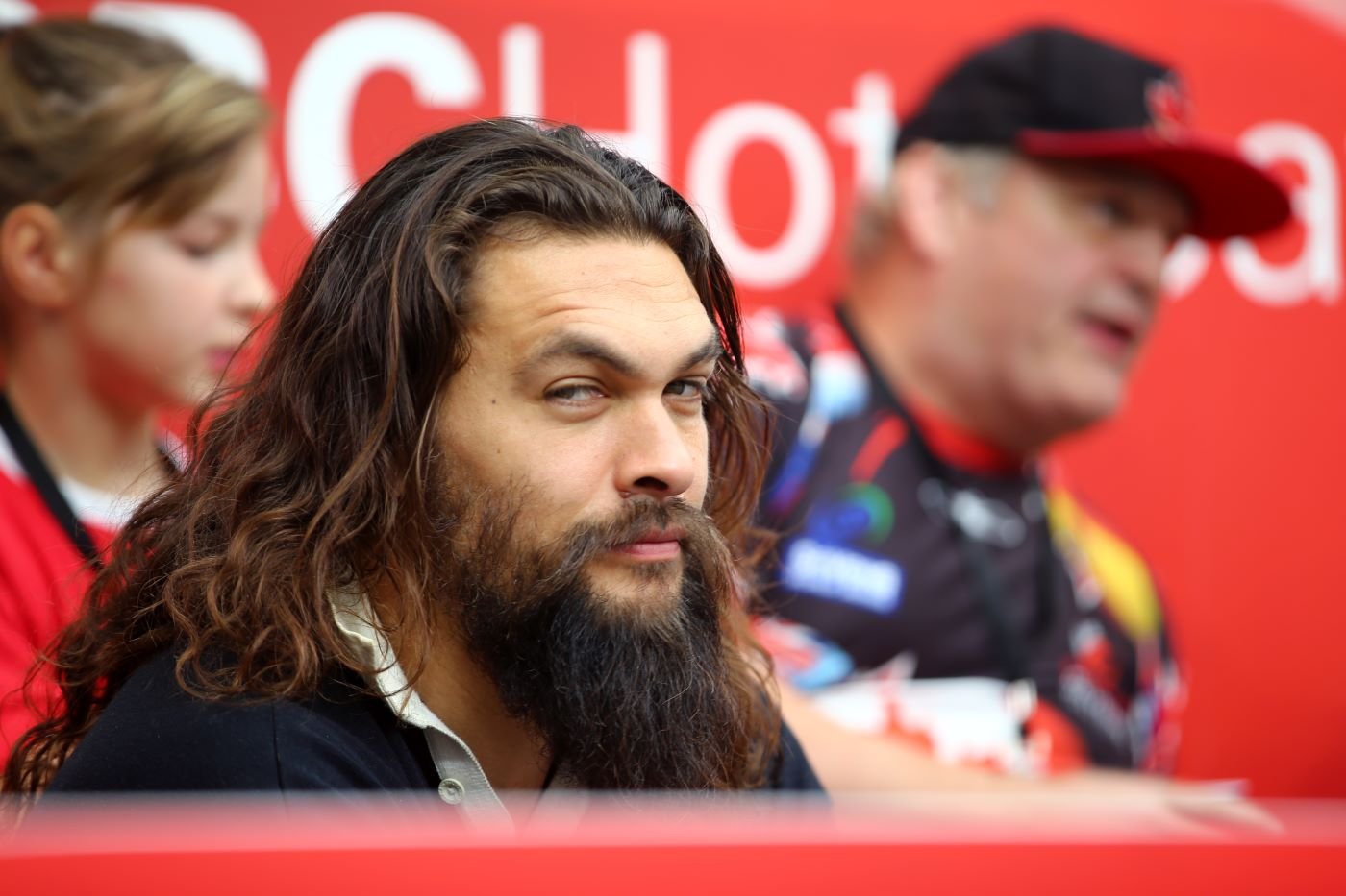 Jason Momoa wearing a black shirt with a white collar against a red background with a couple people blurred out in the background.