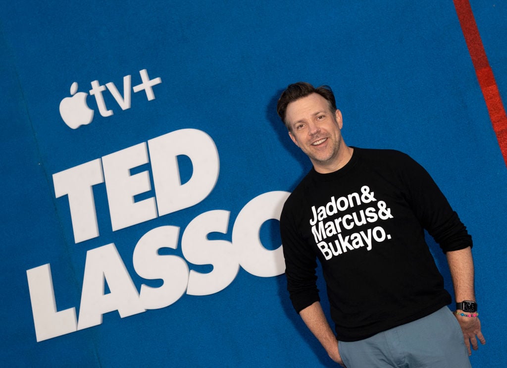 Jason Sudeikis walks the red carpet while wearing a black t-shirt. A 'Ted Lasso' poster is behind him.