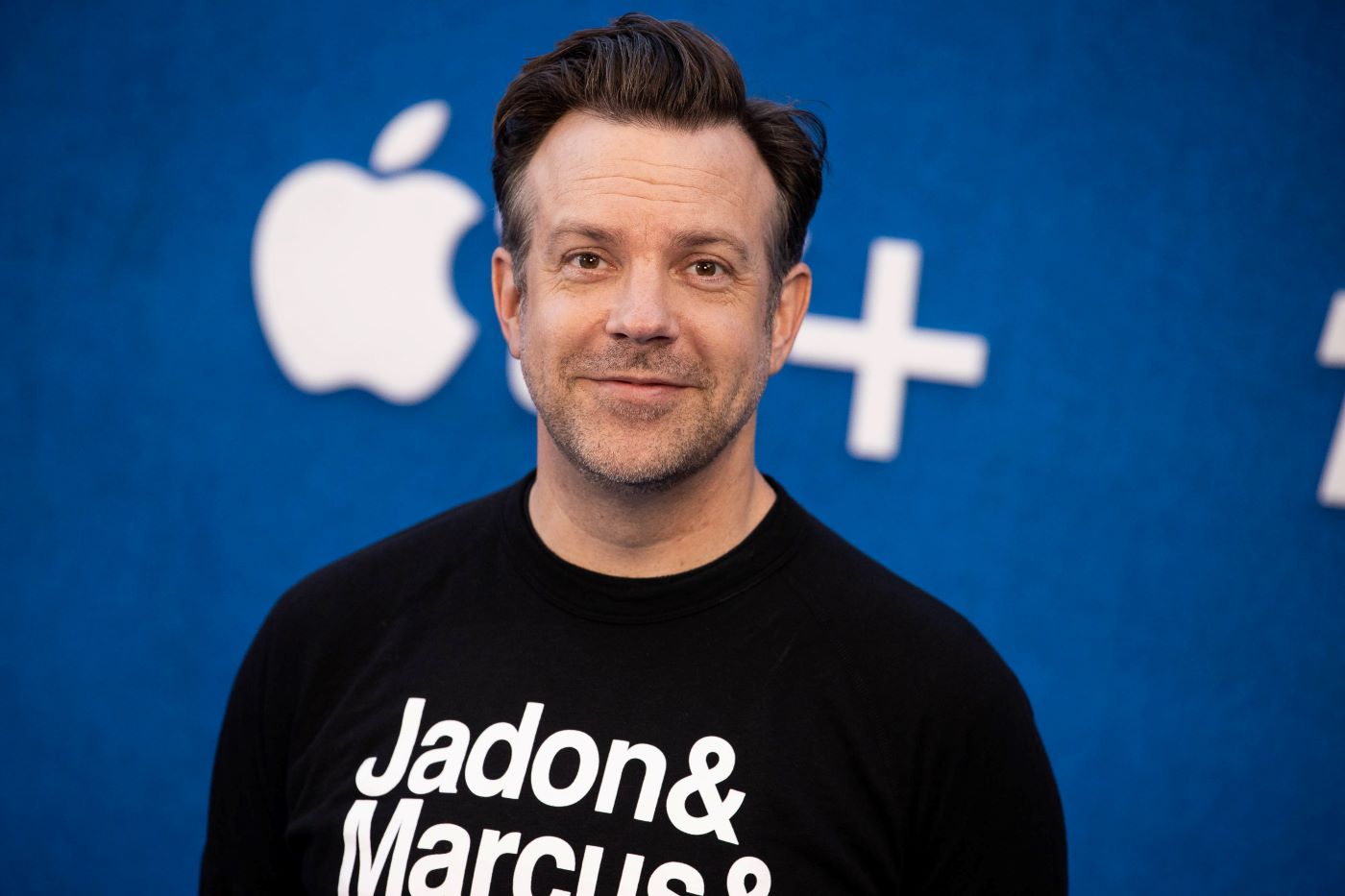 Jason Sudeikis from 'Ted Lasso' wearing a black shirt with white writing standing in front of a blue background with white writing.