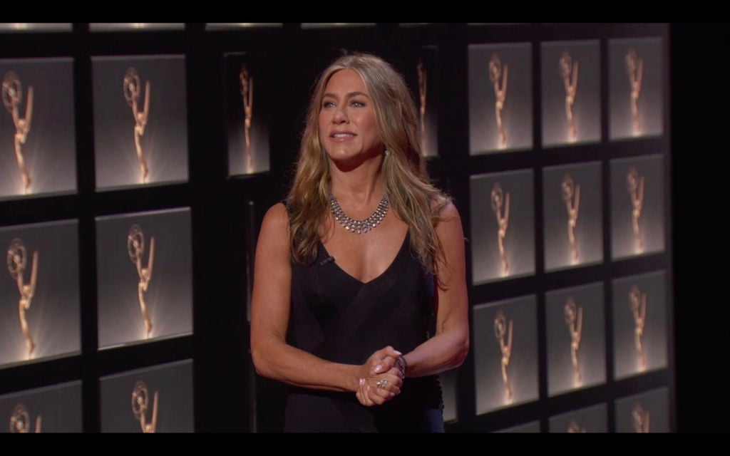 Jennifer Aniston stands backstage at the Emmy Awards. She's wearing a black dress with a large silver necklace and her golden hair is down.