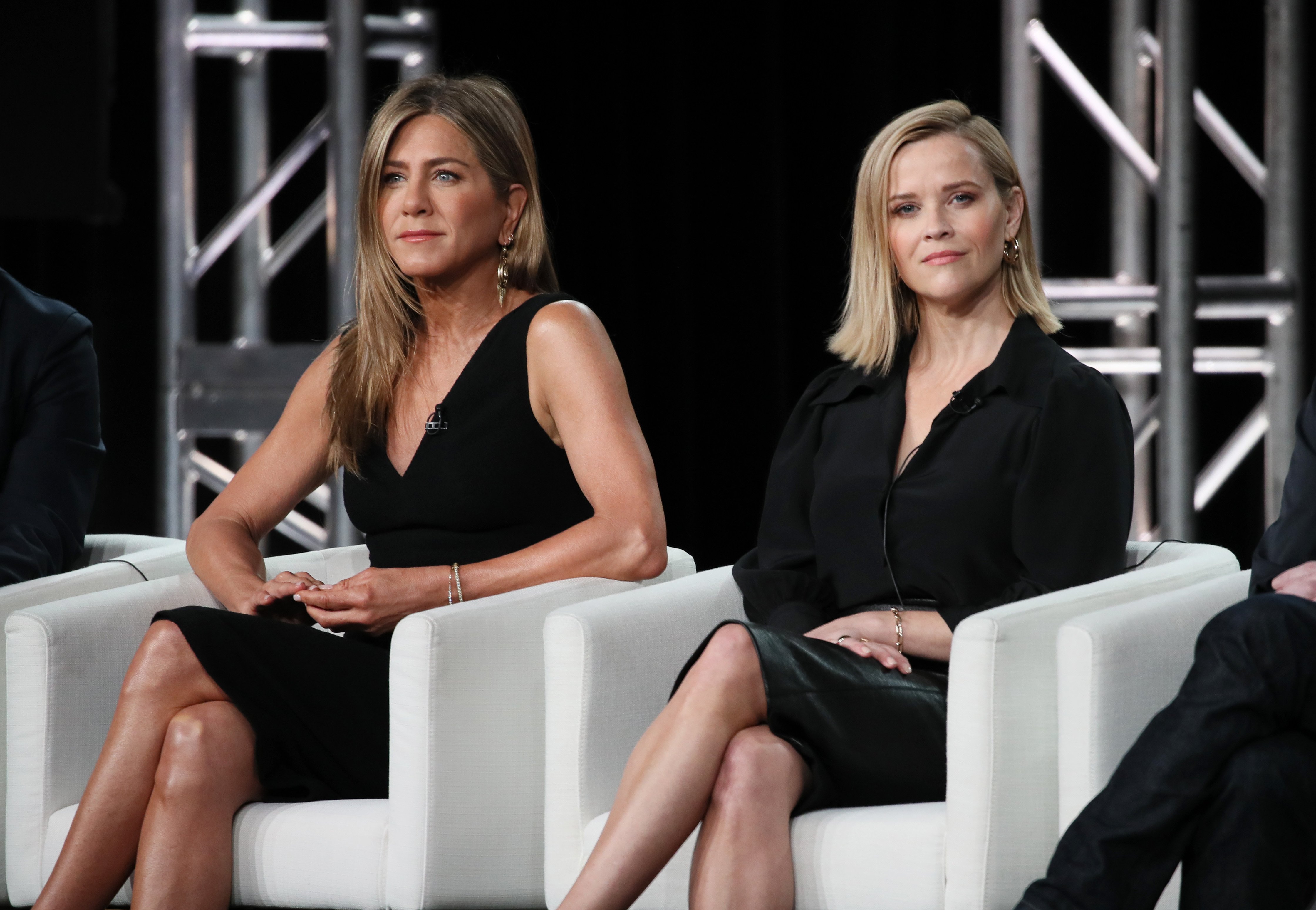 Jennifer Aniston and Reese Witherspoon sit side by side in white chairs wearing black dresses.