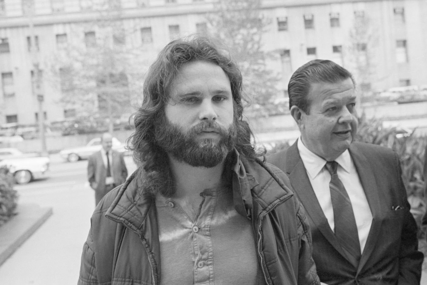 Jim Morrison, member of the 'Doors' standing in front of a building with many windows in a black and white picture.