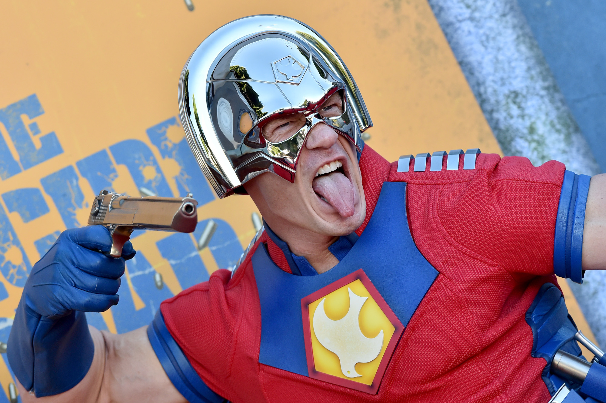 John Cena dressed as Peacemaker at 'The Suicide Squad' premiere. He's wearing a silver helmet and red, yellow, and blue spandex shirt. He's sticking his tongue out and holding a fake gun up.