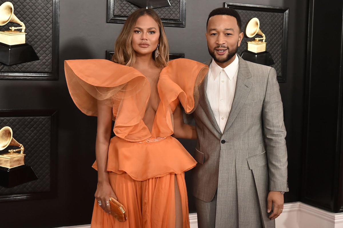 Chrissy Teigen wears an orange gown and poses with husband John Legend, who wears a gray tuxedo.