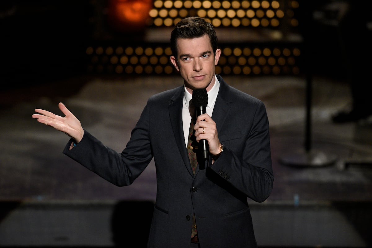 John Mulaney speaks into a microphone on stage.