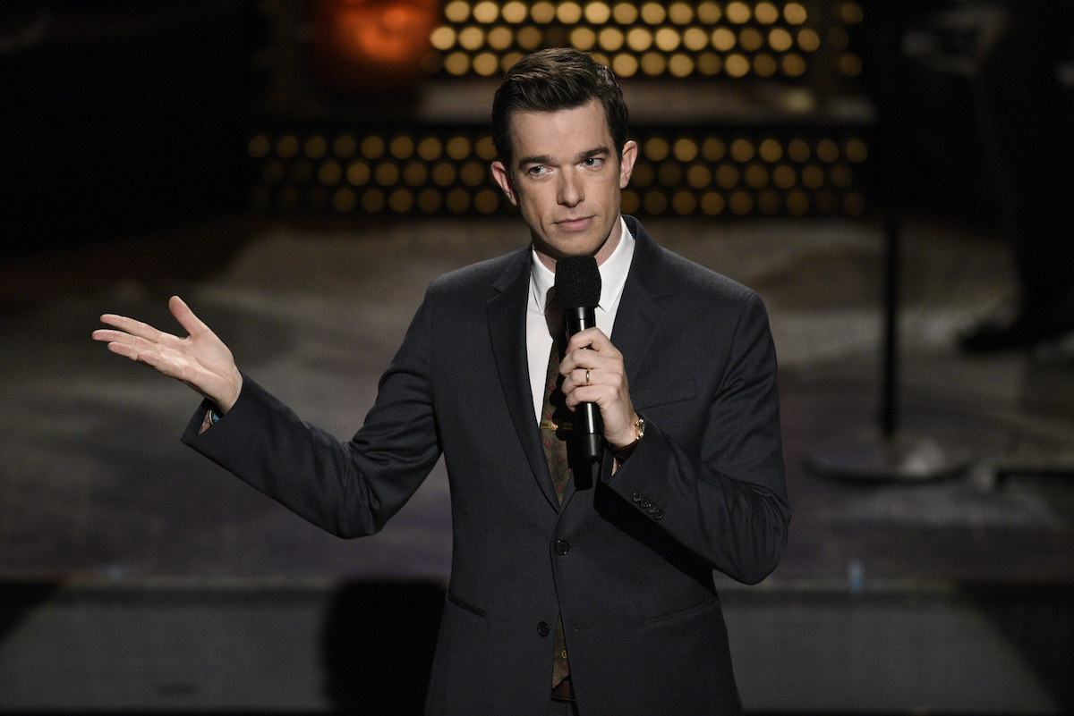John Mulaney holds a microphone and makes hand gestures on stage.