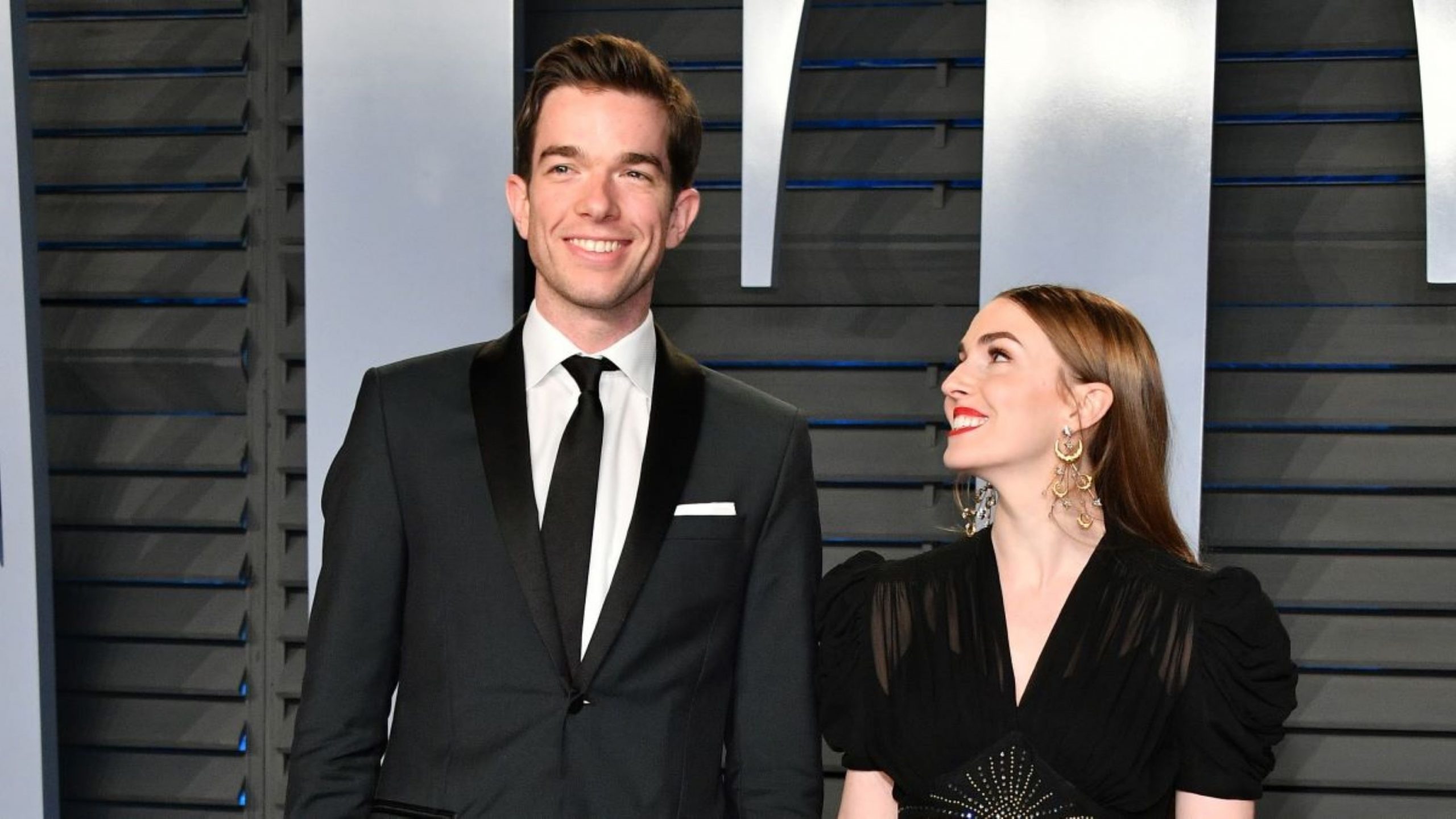 John Mulaney in black suit smiling, and Anna Marie Tendler smiling up at him