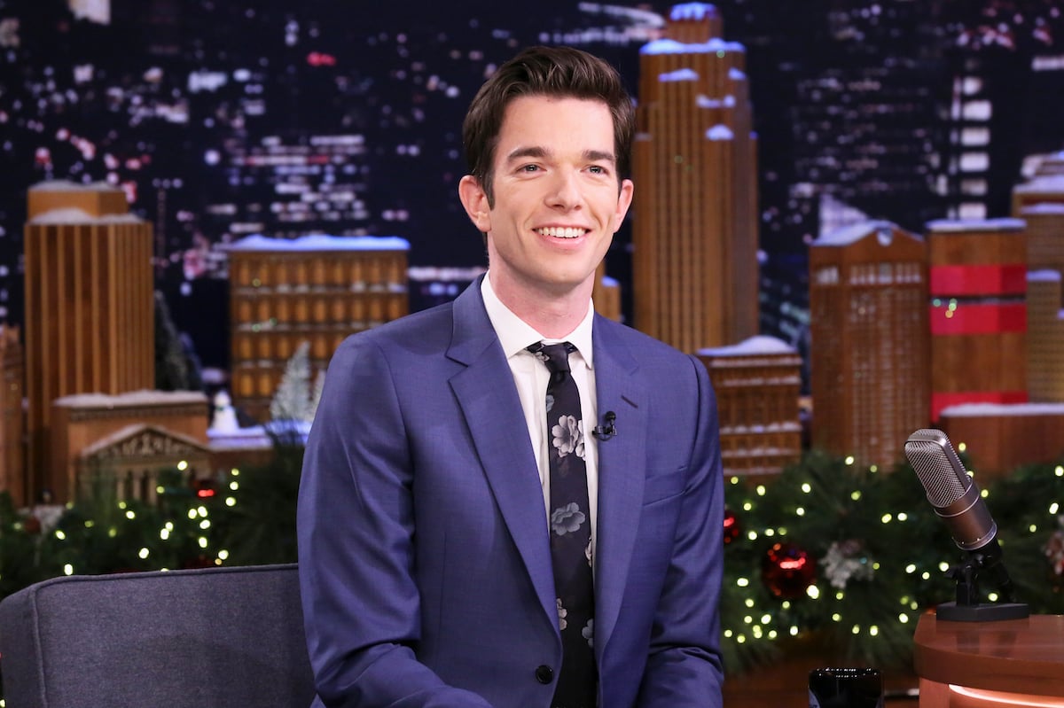John Mulaney wearing a blue suit and floral tie and smiling for the camera.